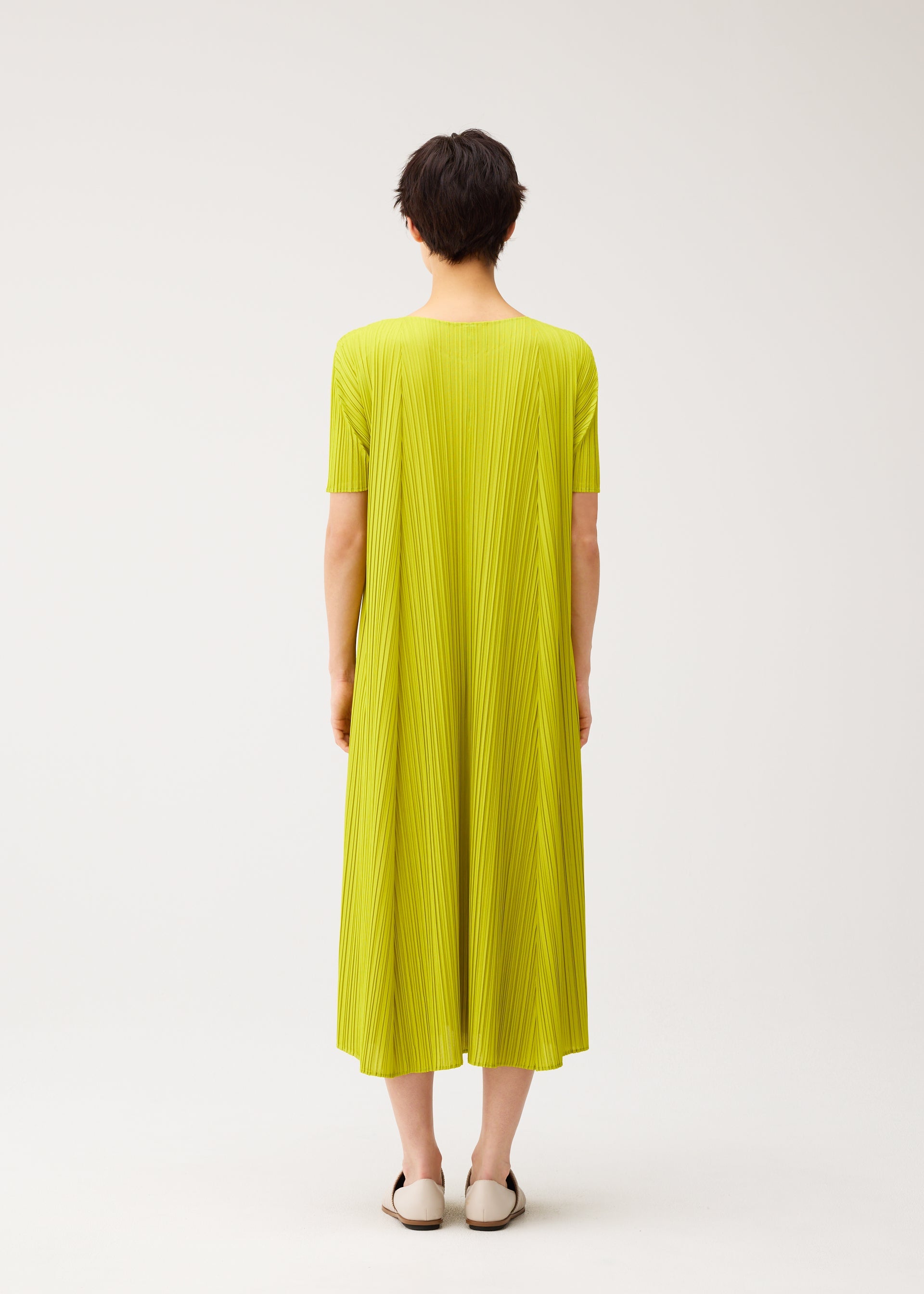 Zoomed view A person with short dark hair faces away, wearing the MELLOW PLEATS dress by PLEATS PLEASE ISSEY MIYAKE in bright yellow-green with a round neck and pleated short sleeves, paired with white slip-on shoes against a plain background.