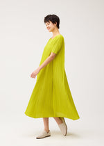 A person with short dark hair smiles while wearing the PLEATS PLEASE ISSEY MIYAKE MELLOW PLEATS bright yellow short-sleeved dress and light beige slip-on shoes against a plain white background.