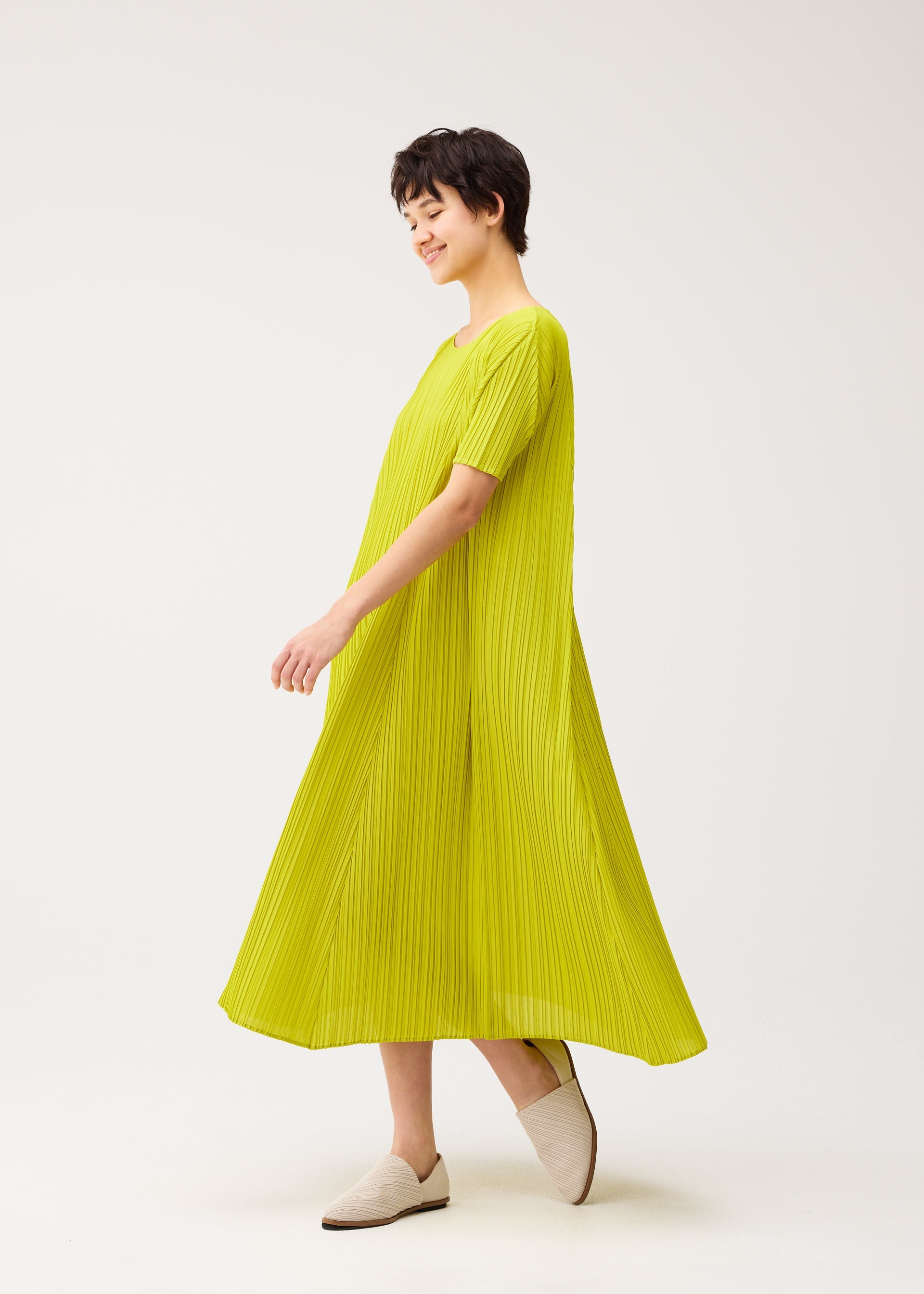 Zoomed view A person with short dark hair smiles while wearing the PLEATS PLEASE ISSEY MIYAKE MELLOW PLEATS bright yellow short-sleeved dress and light beige slip-on shoes against a plain white background.
