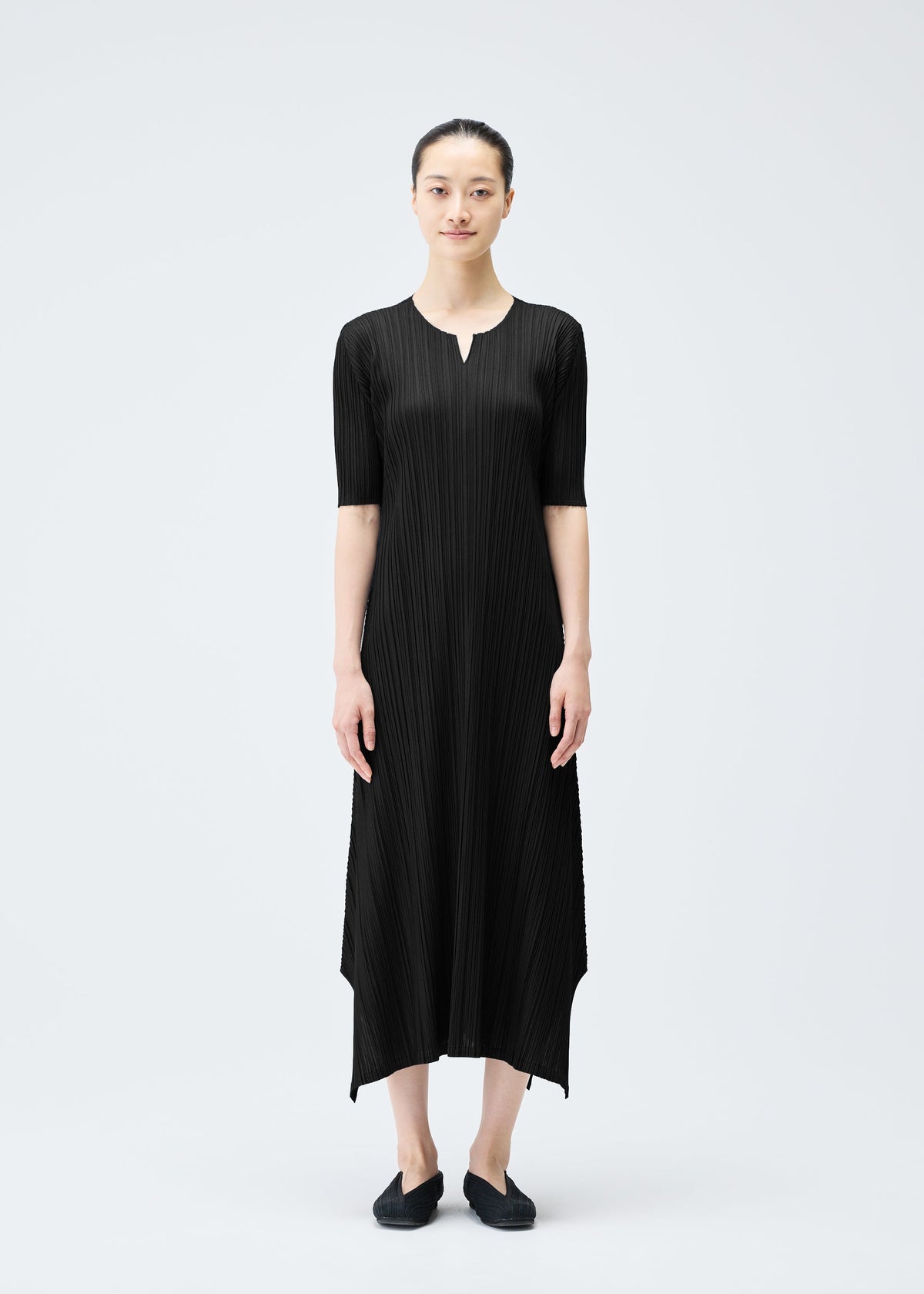 A woman stands against a plain light background wearing the MELLOW PLEATS ankle-length black dress by PLEATS PLEASE ISSEY MIYAKE, featuring pleats, elbow sleeves, a notched neckline, and paired with black flats and her hair pulled back. view 6 of 9