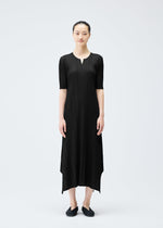A woman stands against a plain light background wearing the MELLOW PLEATS ankle-length black dress by PLEATS PLEASE ISSEY MIYAKE, featuring pleats, elbow sleeves, a notched neckline, and paired with black flats and her hair pulled back.