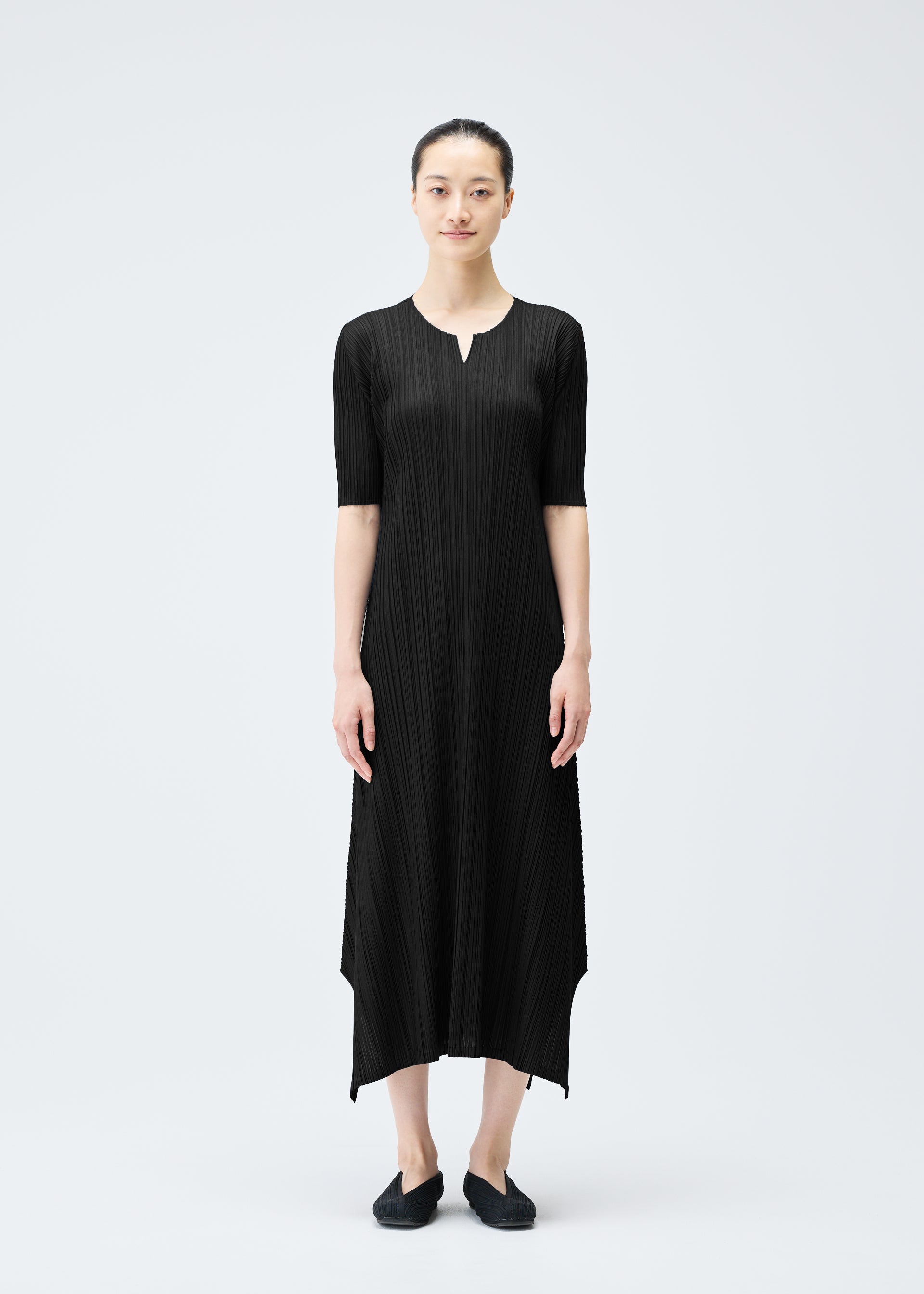 Zoomed view A woman stands against a plain light background wearing the MELLOW PLEATS ankle-length black dress by PLEATS PLEASE ISSEY MIYAKE, featuring pleats, elbow sleeves, a notched neckline, and paired with black flats and her hair pulled back.