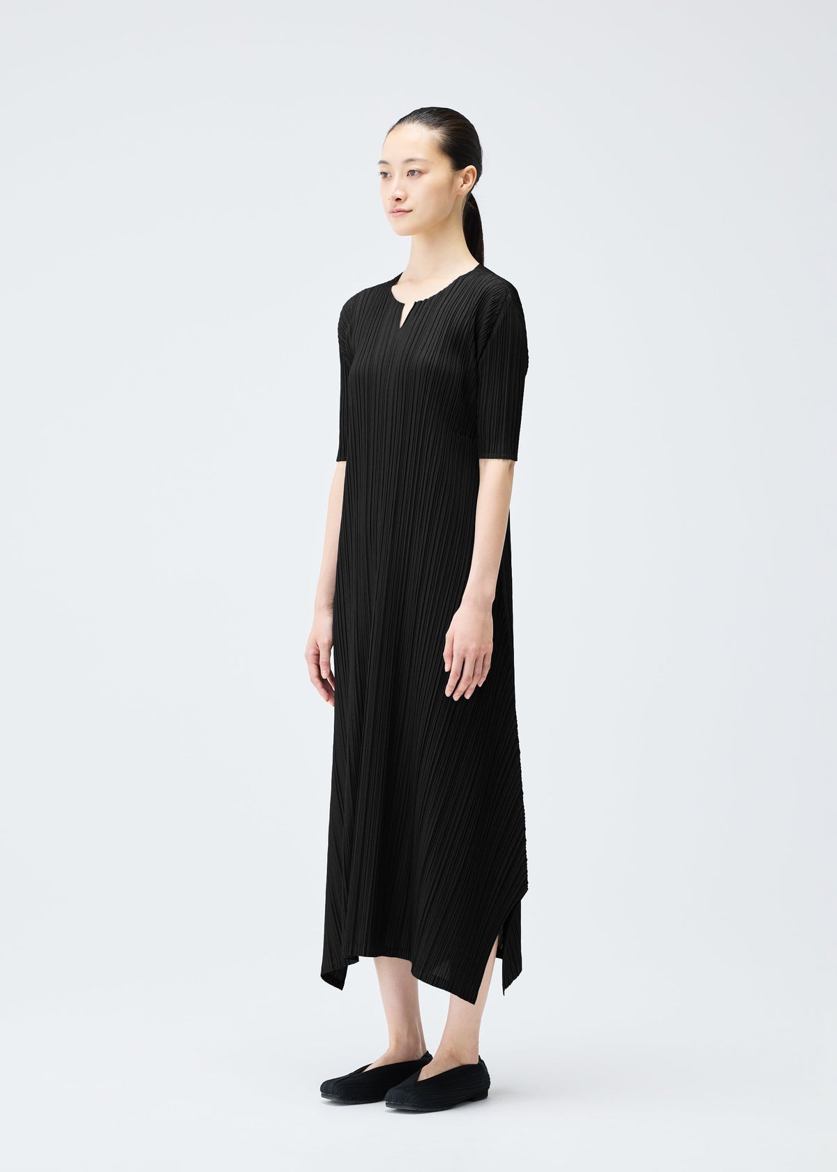 A woman with dark hair in a low ponytail stands against a white background, wearing the black MELLOW PLEATS ankle-length V-neck dress by PLEATS PLEASE ISSEY MIYAKE and black slip-on shoes. view 7 of 9