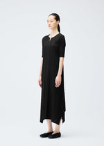A woman with dark hair in a low ponytail stands against a white background, wearing the black MELLOW PLEATS ankle-length V-neck dress by PLEATS PLEASE ISSEY MIYAKE and black slip-on shoes.