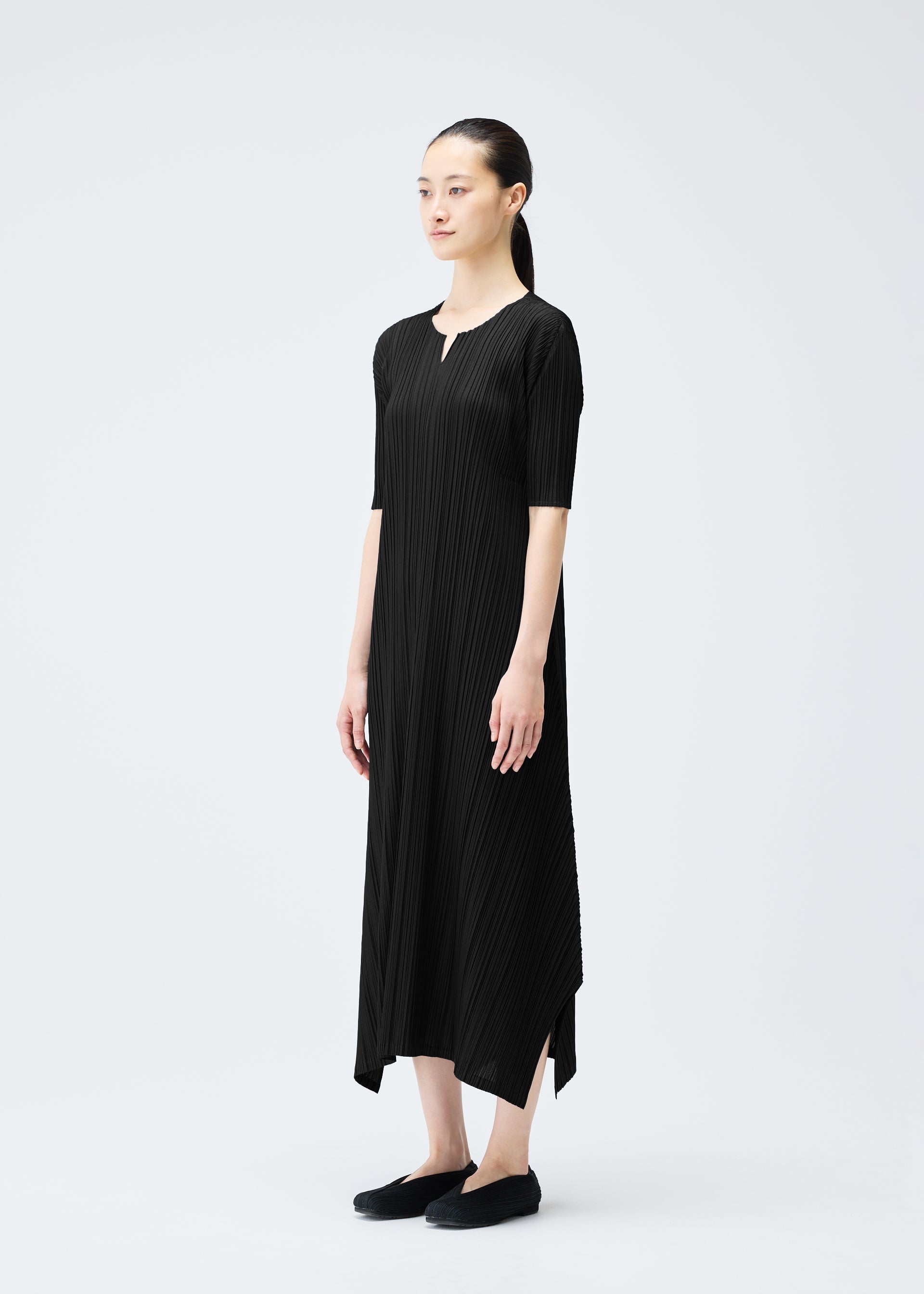 Zoomed view A woman with dark hair in a low ponytail stands against a white background, wearing the black MELLOW PLEATS ankle-length V-neck dress by PLEATS PLEASE ISSEY MIYAKE and black slip-on shoes.