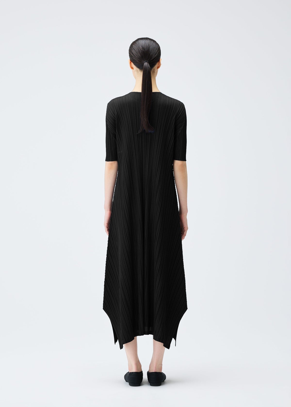 A woman with straight dark hair in a low ponytail stands facing away in the black, pleated MELLOW PLEATS dress by PLEATS PLEASE ISSEY MIYAKE, paired with black shoes, against a plain white background. view 8 of 9