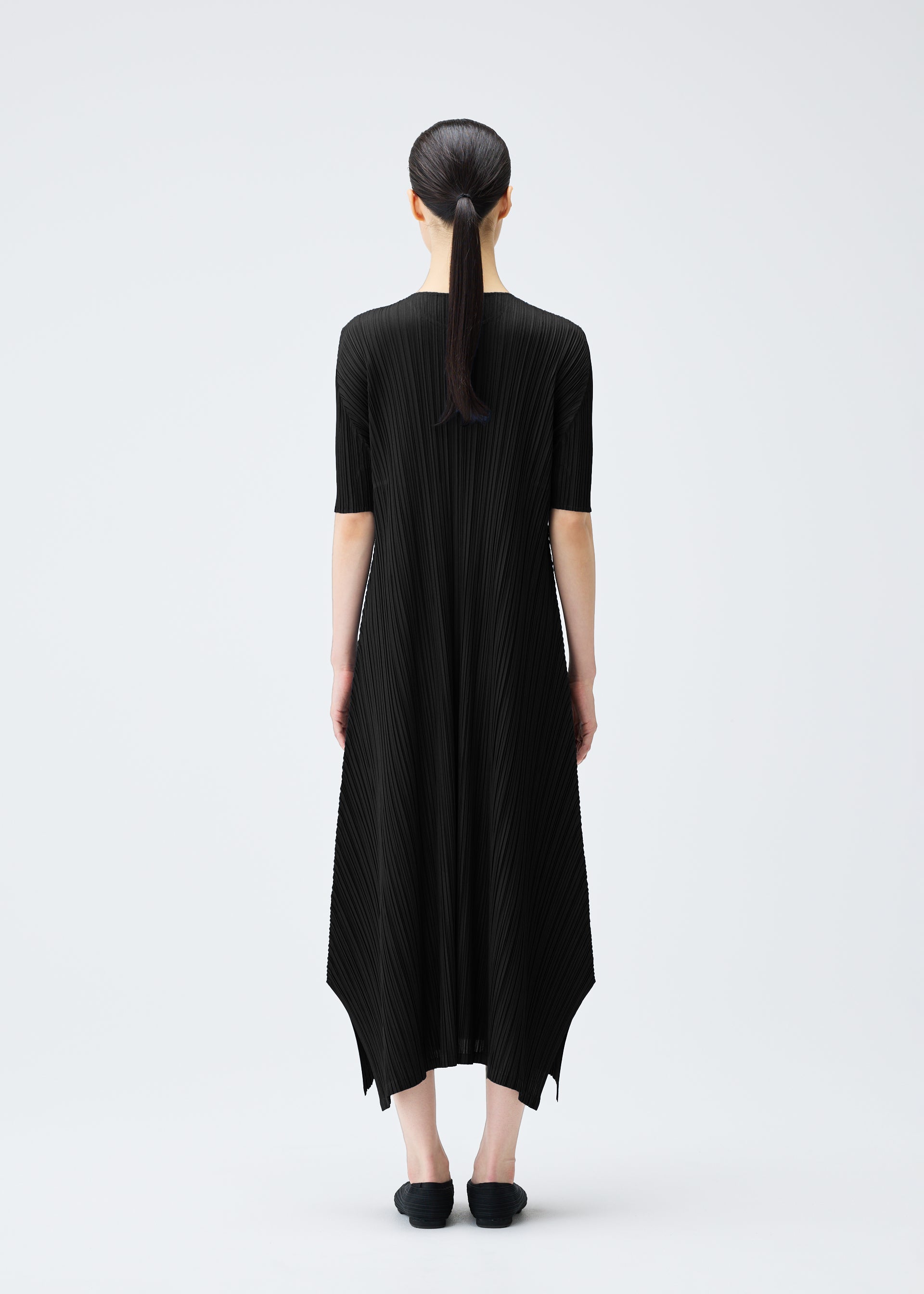 Zoomed view A woman with straight dark hair in a low ponytail stands facing away in the black, pleated MELLOW PLEATS dress by PLEATS PLEASE ISSEY MIYAKE, paired with black shoes, against a plain white background.