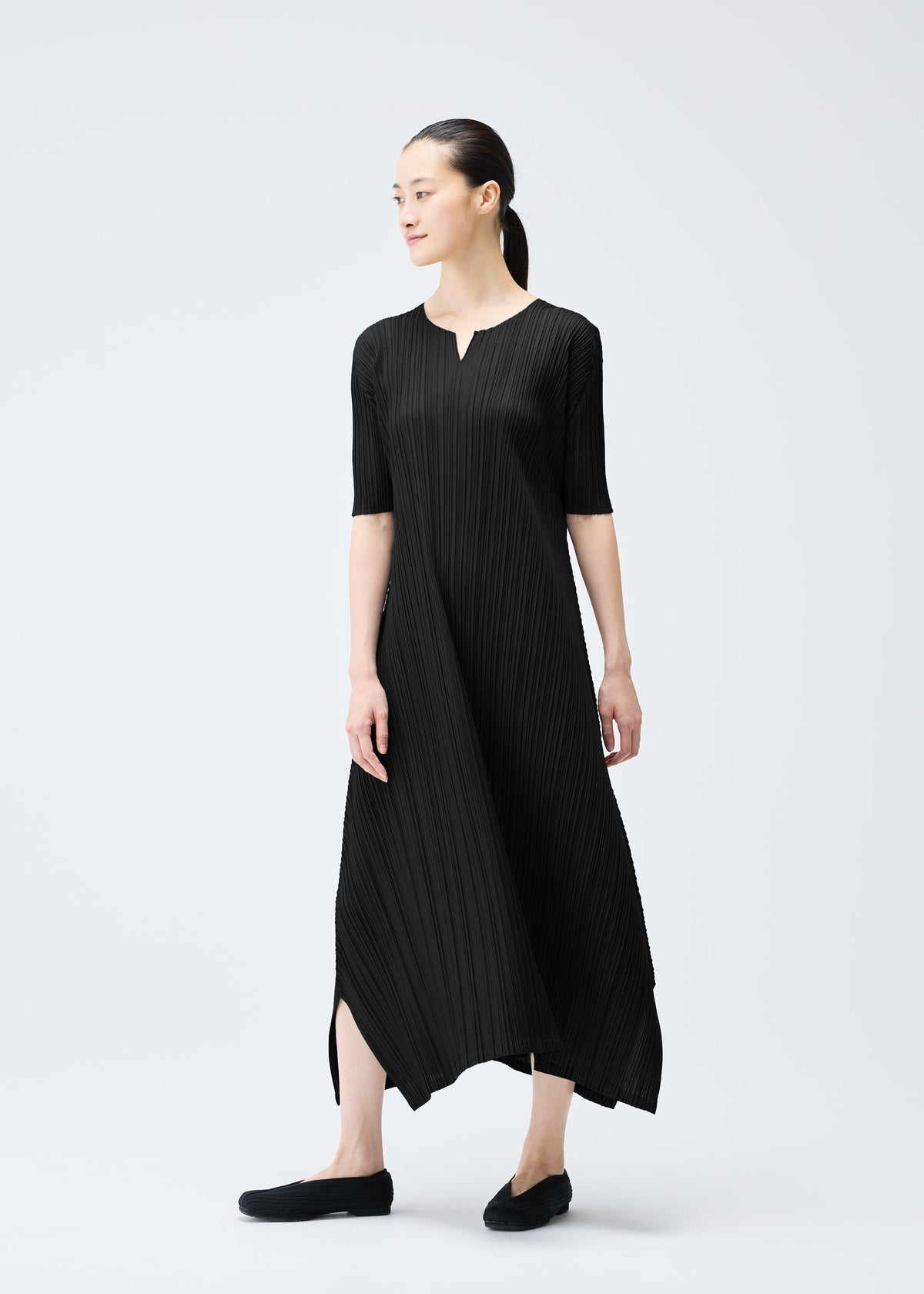 A woman stands against a plain light background, wearing the MELLOW PLEATS dress by PLEATS PLEASE ISSEY MIYAKE—black, mid-calf length, elbow sleeves, small V-neck. She looks to the side and pairs it with black shoes. view 9 of 9