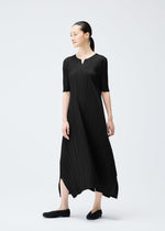 A woman stands against a plain light background, wearing the MELLOW PLEATS dress by PLEATS PLEASE ISSEY MIYAKE—black, mid-calf length, elbow sleeves, small V-neck. She looks to the side and pairs it with black shoes.