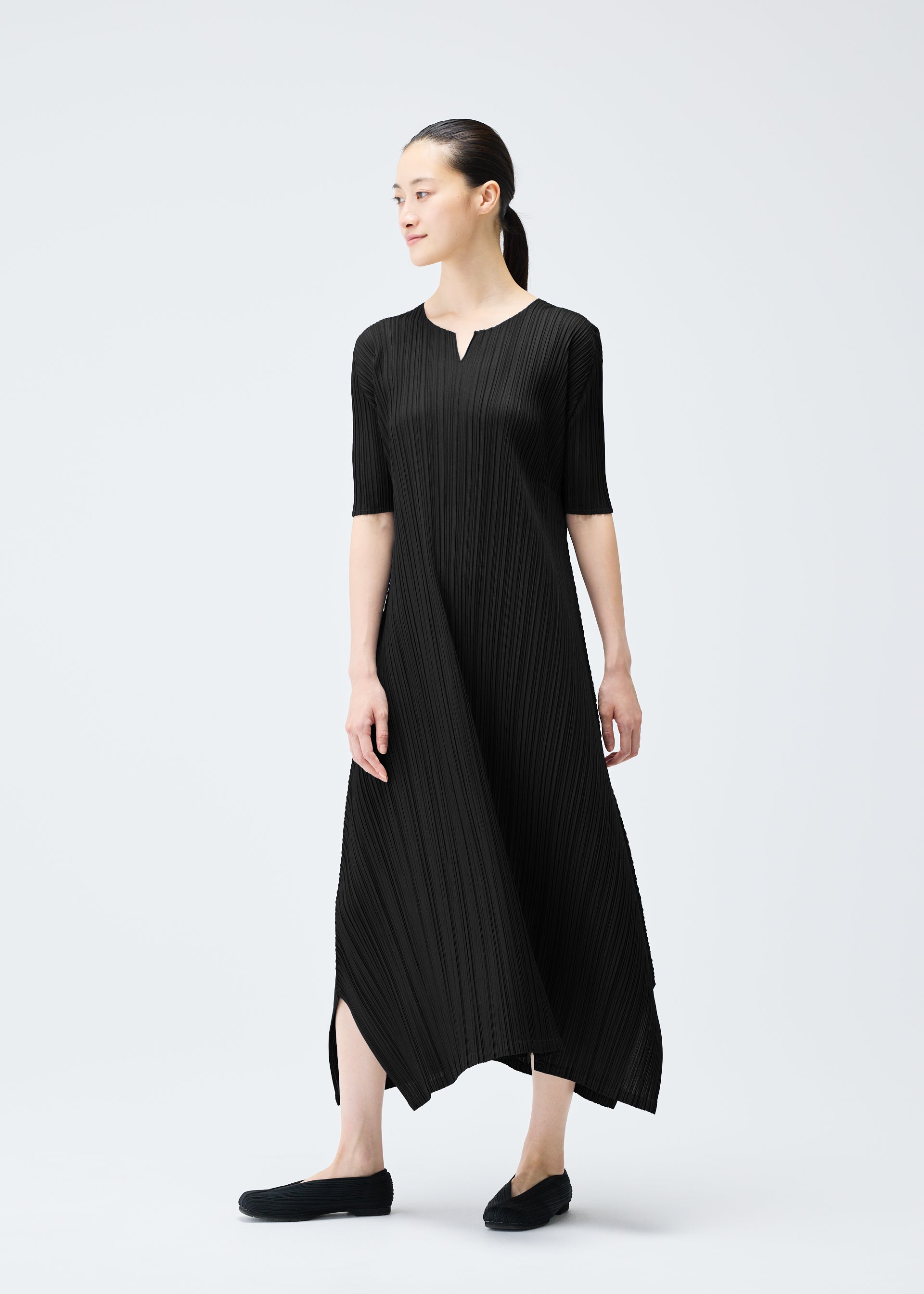 Zoomed view A woman stands against a plain light background, wearing the MELLOW PLEATS dress by PLEATS PLEASE ISSEY MIYAKE—black, mid-calf length, elbow sleeves, small V-neck. She looks to the side and pairs it with black shoes.