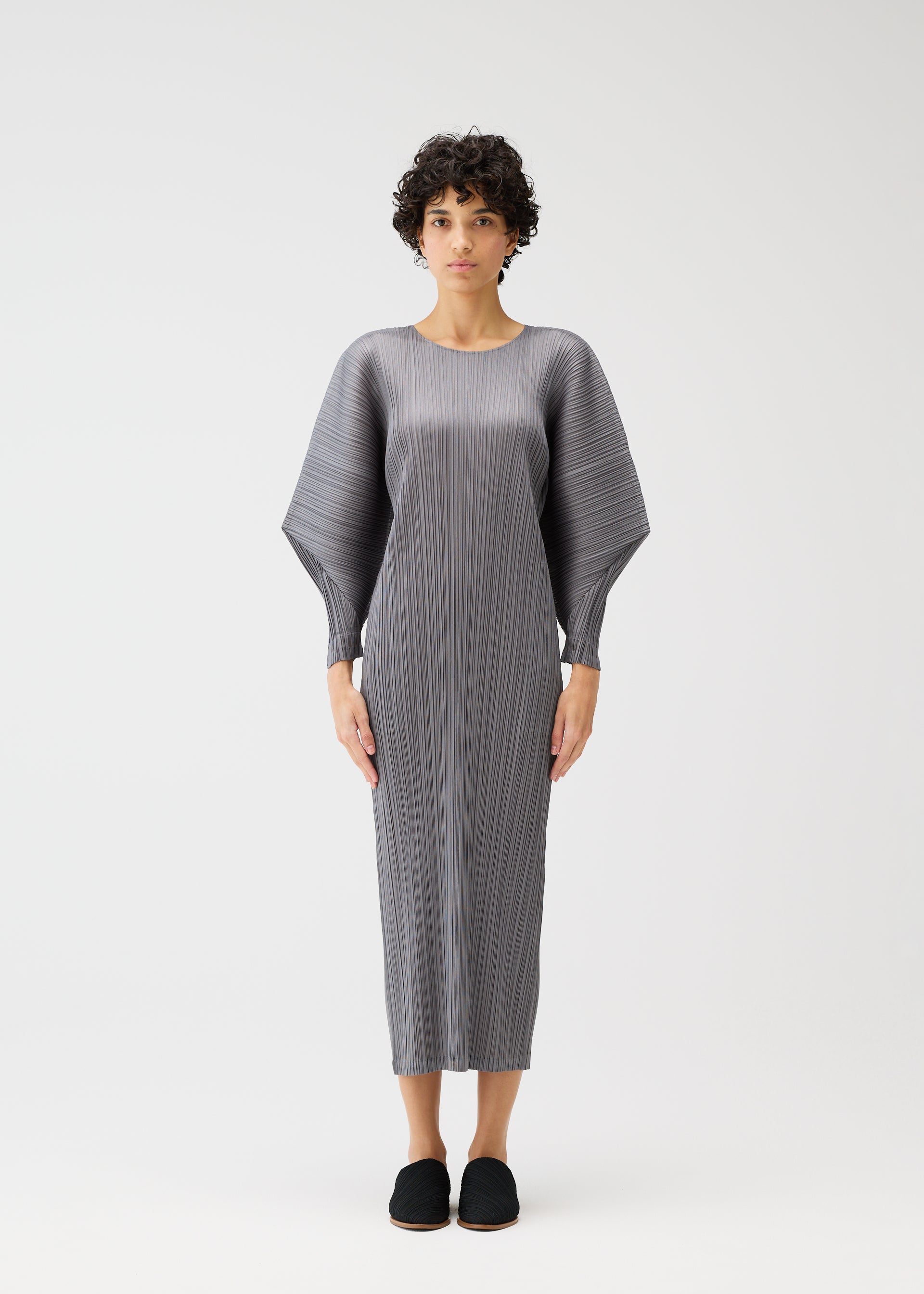 Zoomed view A person with short curly hair models the long, gray SKYLINE pleated round-neck dress by PLEATS PLEASE ISSEY MIYAKE, featuring wide structured dolman sleeves, standing against a plain white background facing forward.