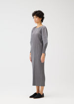 A person with short curly hair stands sideways, wearing the SKYLINE long gray pleated dress with round neck and dolman sleeves from PLEATS PLEASE ISSEY MIYAKE, paired with black slip-on shoes, against a plain white background.