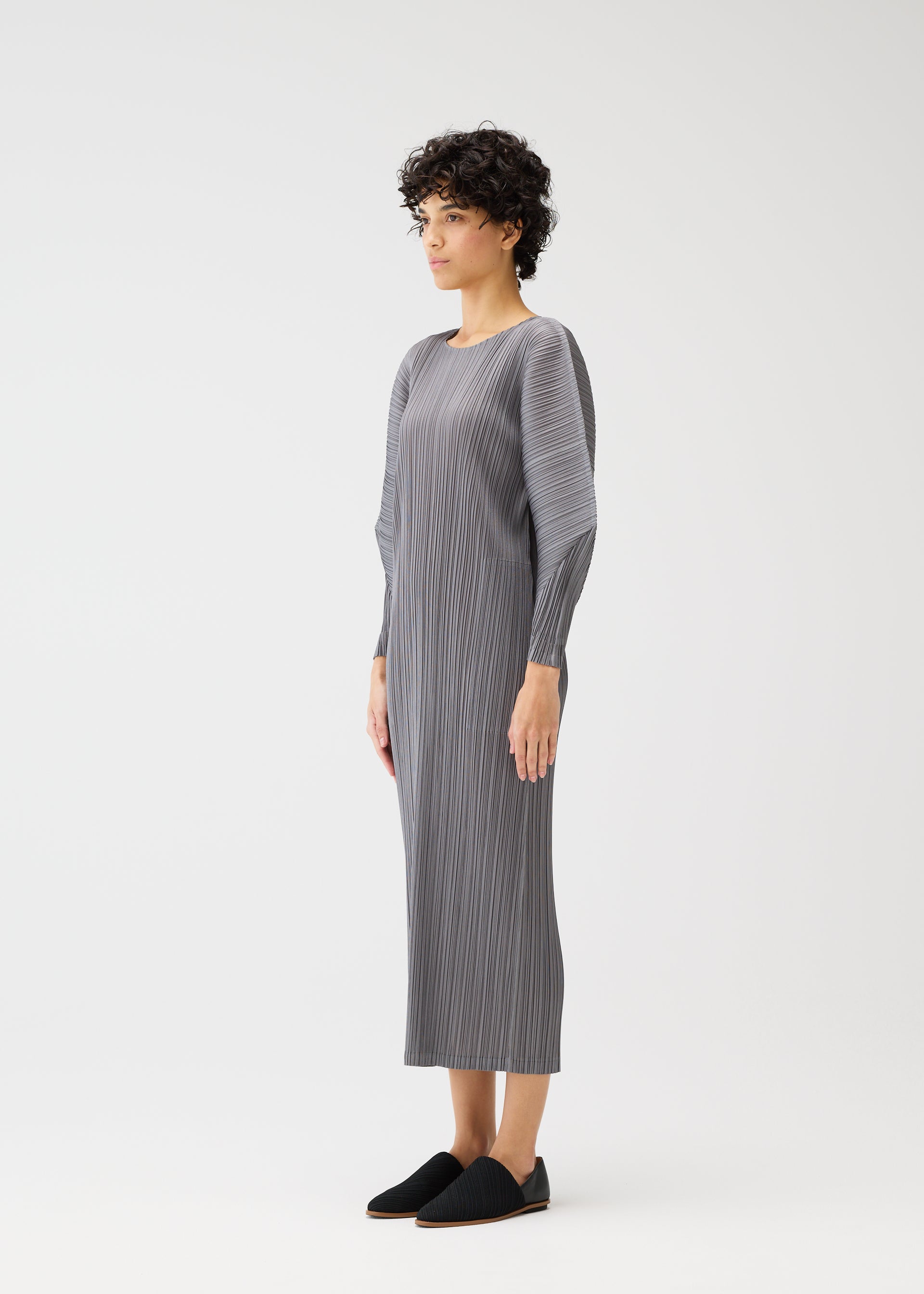 Zoomed view A person with short curly hair stands sideways, wearing the SKYLINE long gray pleated dress with round neck and dolman sleeves from PLEATS PLEASE ISSEY MIYAKE, paired with black slip-on shoes, against a plain white background.