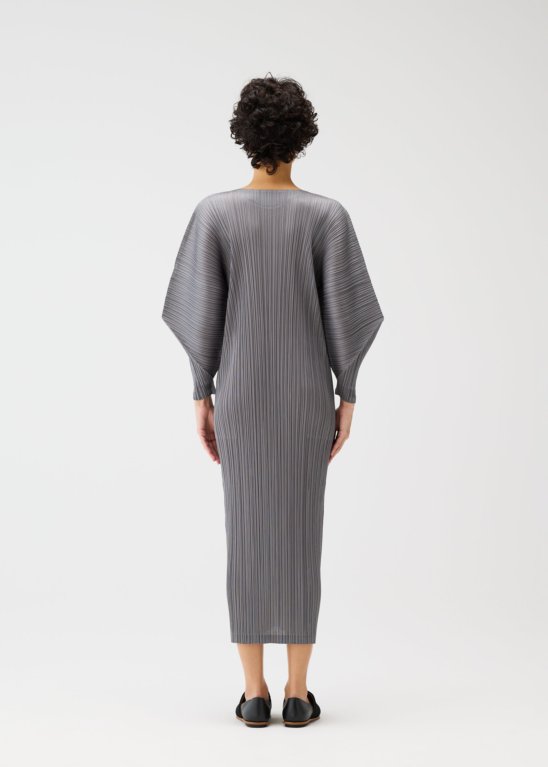 Zoomed view A person with short, curly hair is seen from behind wearing the SKYLINE long gray pleated round-neck dress with dolman sleeves by PLEATS PLEASE ISSEY MIYAKE and black slip-on shoes against a plain white background.