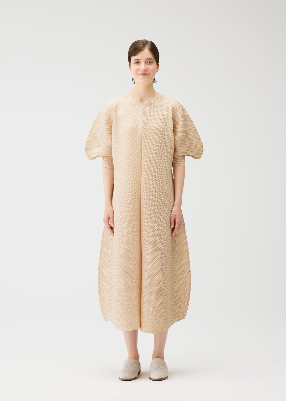 A woman with short brown hair stands against a white background, wearing the COAST dress by PLEATS PLEASE ISSEY MIYAKE—a beige, textured V-neck dress with pleats, wave patterns, voluminous short sleeves, and a loose ankle-length skirt. view 5 of 8