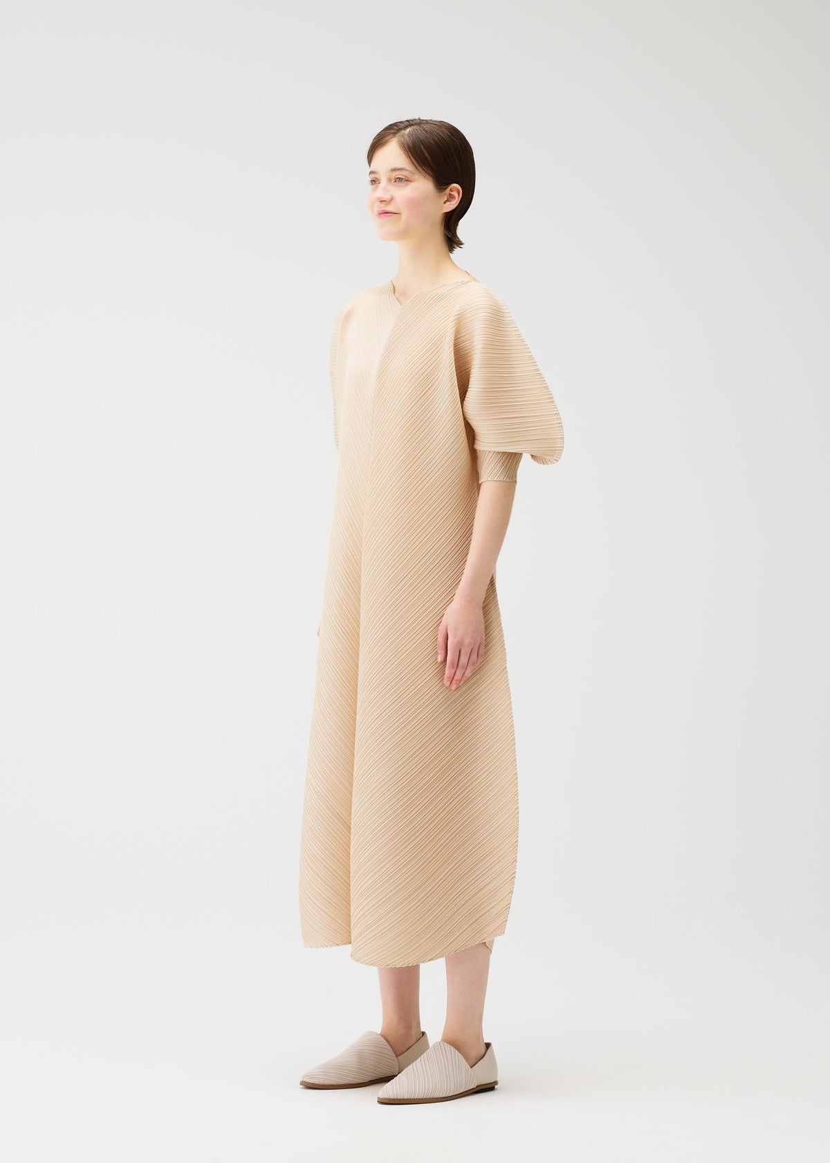 A person wears the COAST dress by PLEATS PLEASE ISSEY MIYAKE—a light beige, wave pattern V-neck style with elbow-length balloon sleeves—paired with matching slip-on shoes, standing in a plain white studio and looking slightly left. view 6 of 8