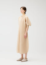 A person wears the COAST dress by PLEATS PLEASE ISSEY MIYAKE—a light beige, wave pattern V-neck style with elbow-length balloon sleeves—paired with matching slip-on shoes, standing in a plain white studio and looking slightly left.