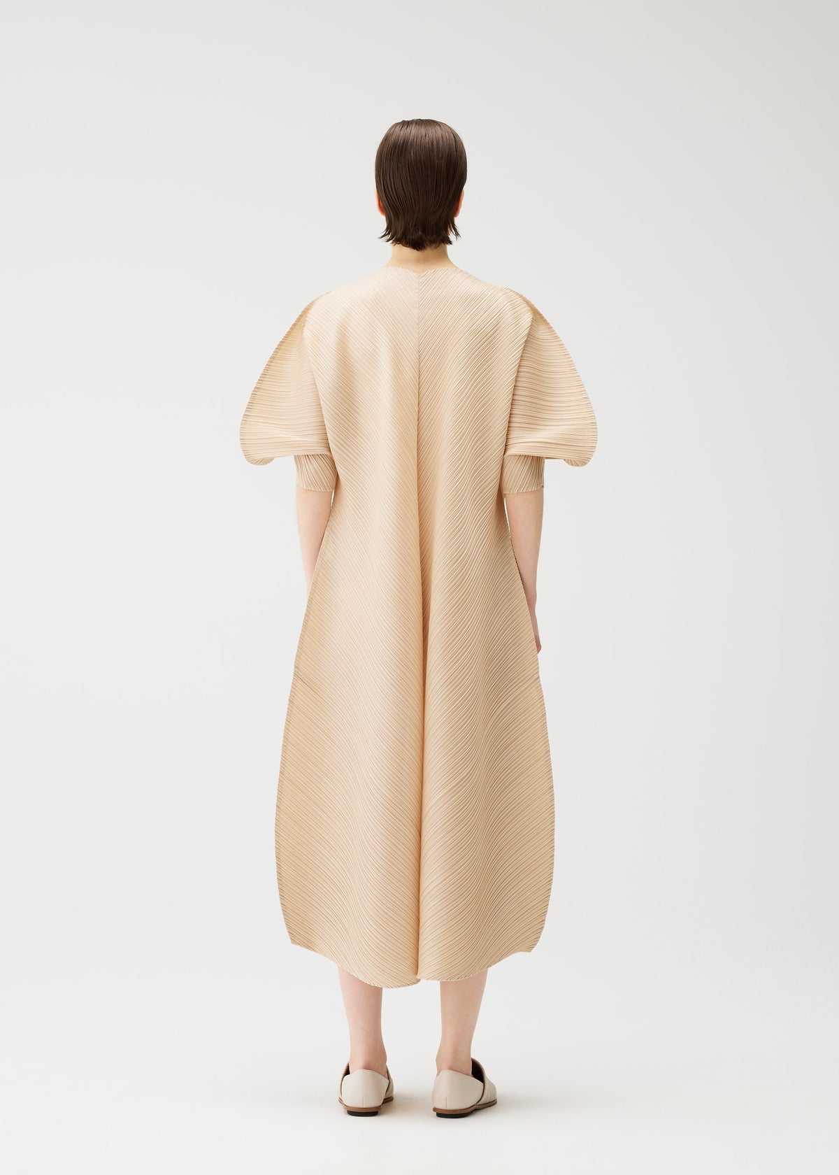 A person with short brown hair stands facing away, wearing the COAST pleated midi V-neck dress with wide sleeves and matching flats by PLEATS PLEASE ISSEY MIYAKE, set against a plain white background. view 7 of 8