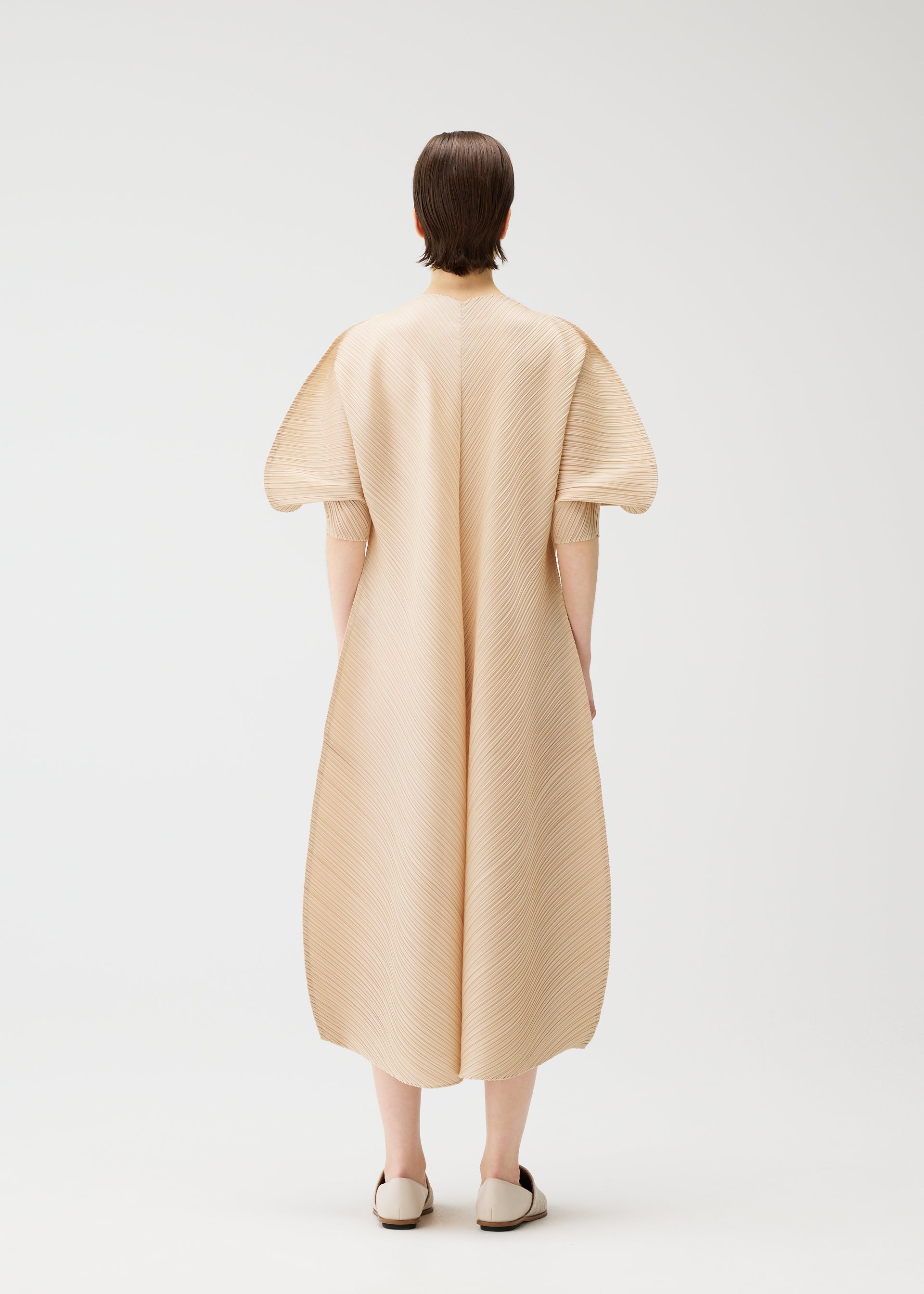 Zoomed view A person with short brown hair stands facing away, wearing the COAST pleated midi V-neck dress with wide sleeves and matching flats by PLEATS PLEASE ISSEY MIYAKE, set against a plain white background.