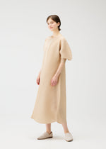 A person with short brown hair and a neutral expression stands against a plain white background, wearing the PLEATS PLEASE ISSEY MIYAKE COAST textured beige V-neck dress with short balloon sleeves and beige slip-on shoes.
