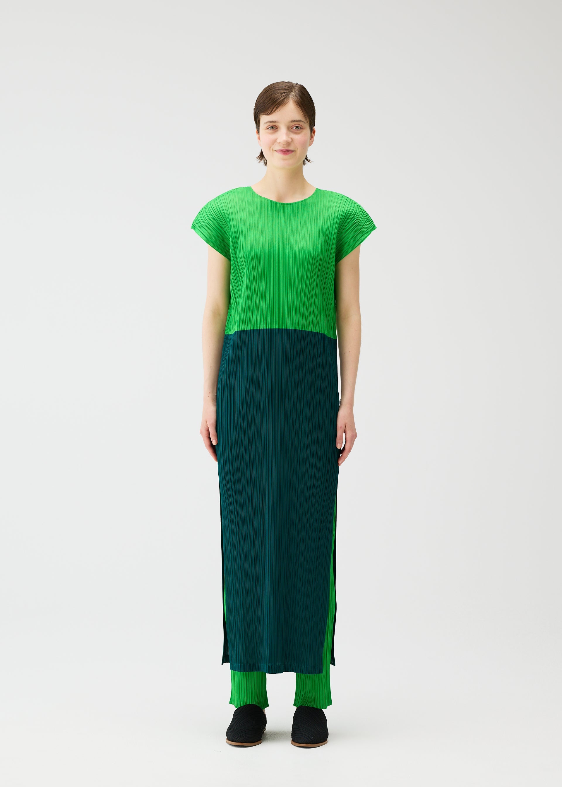 Zoomed view A person stands against a plain white background, wearing the SWING dress by PLEATS PLEASE ISSEY MIYAKE—featuring a two-tone green round-neck design with French sleeves—paired with matching green pants and black shoes.