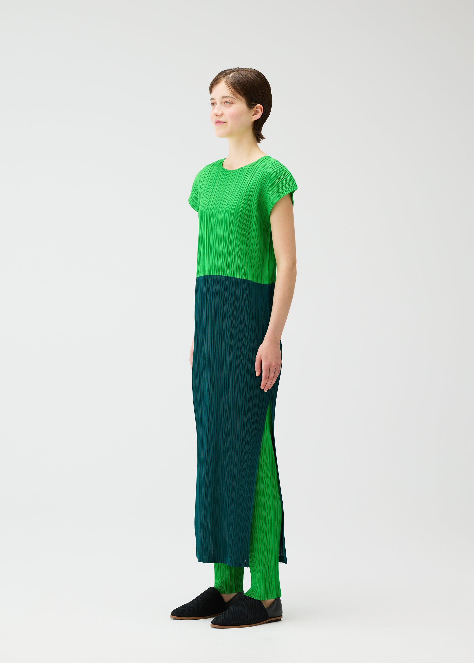Zoomed view A person models the SWING set by PLEATS PLEASE ISSEY MIYAKE—featuring a two-tone round-neck dress with French sleeves and matching pants—paired with black slip-on shoes, standing slightly left on a plain white background.