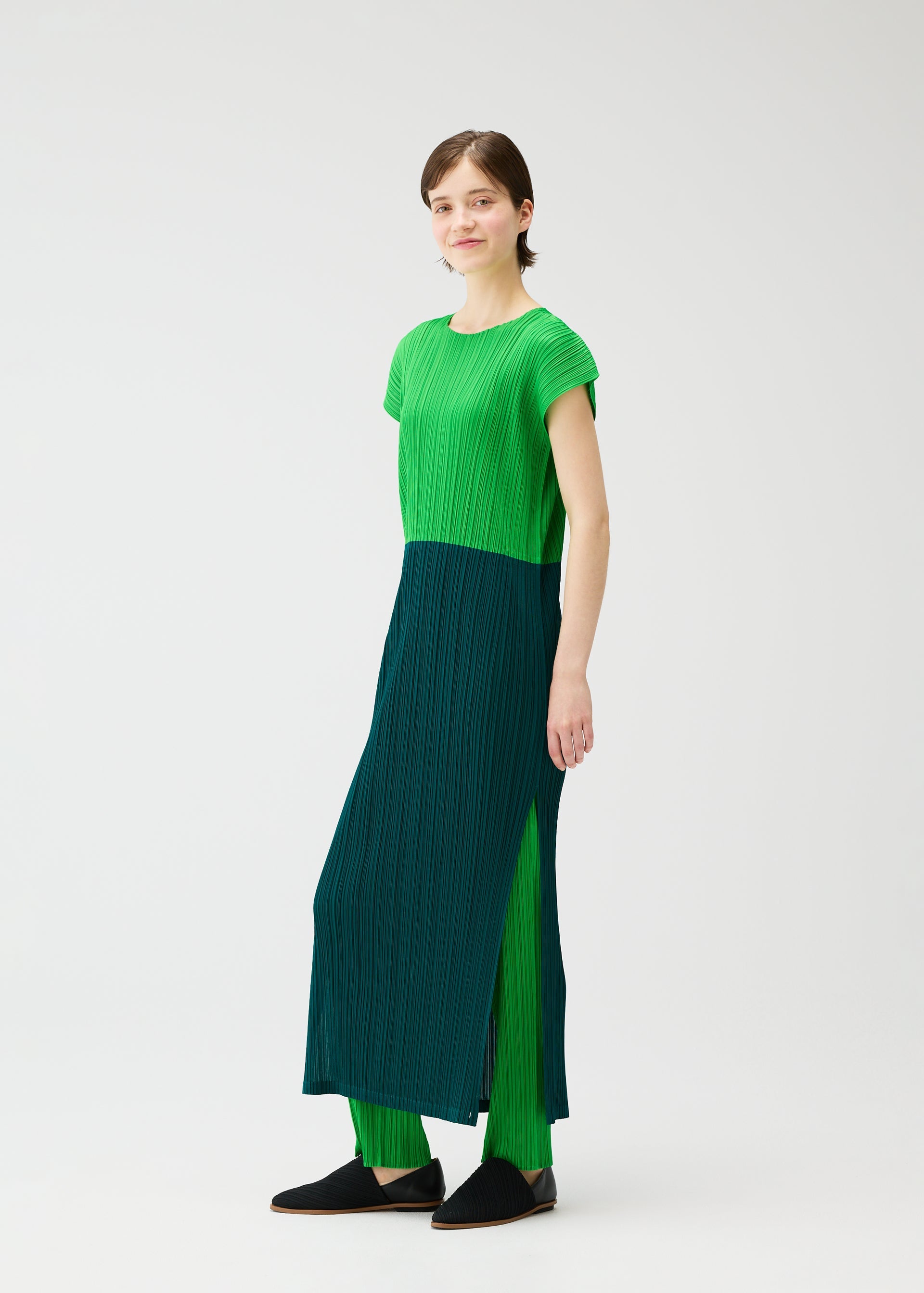 Zoomed view A person with short hair and a neutral expression wears the SWING pleated dress by PLEATS PLEASE ISSEY MIYAKE—featuring a green and dark teal color-block design, French sleeves, matching teal pants, and black slip-on shoes, set against a plain white background.