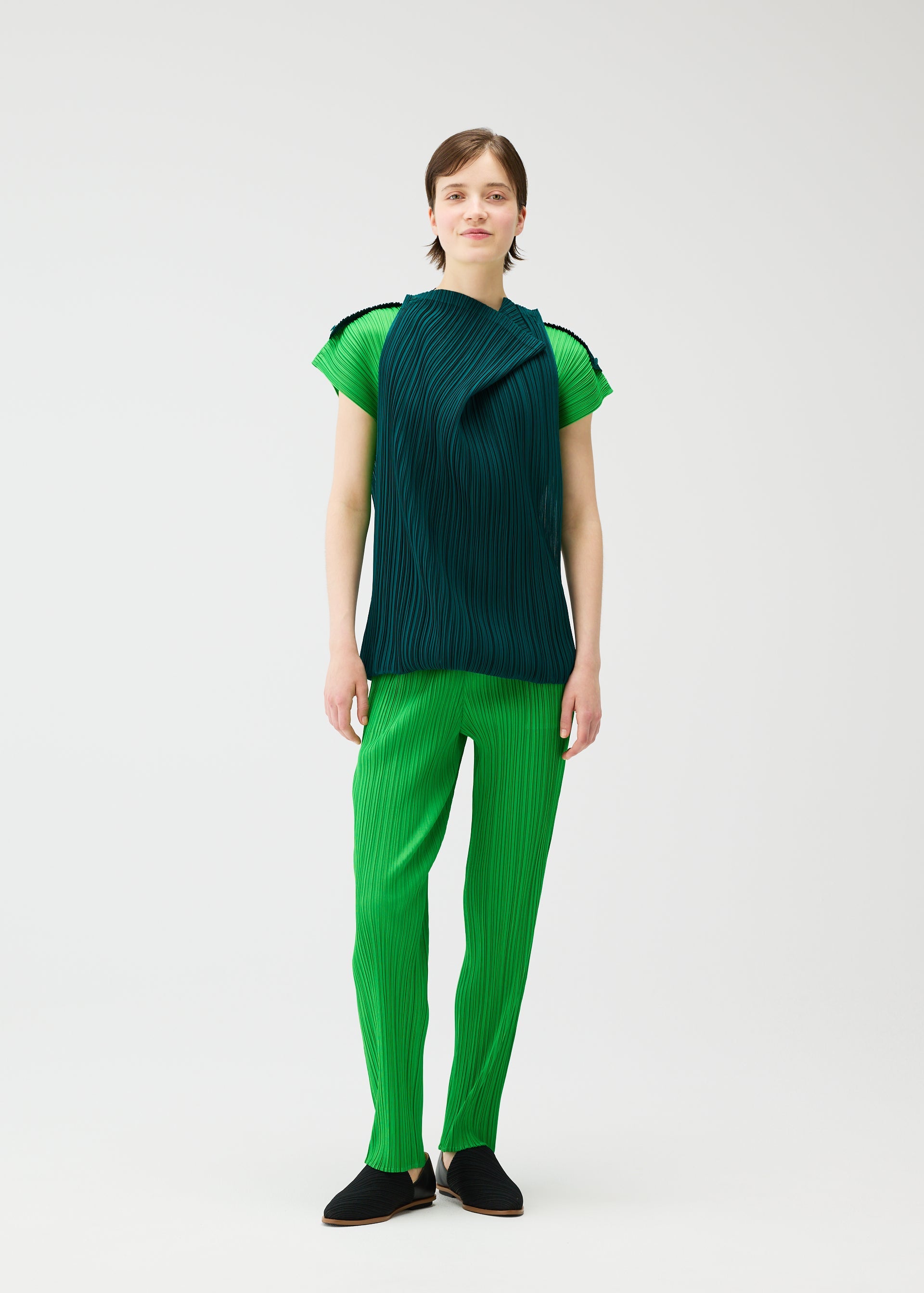 Zoomed view A person stands against a plain white background wearing the SWING top and pants by PLEATS PLEASE ISSEY MIYAKE, featuring a bright green pleated two-tone design with a dark green draped sleeveless overlay and black shoes.