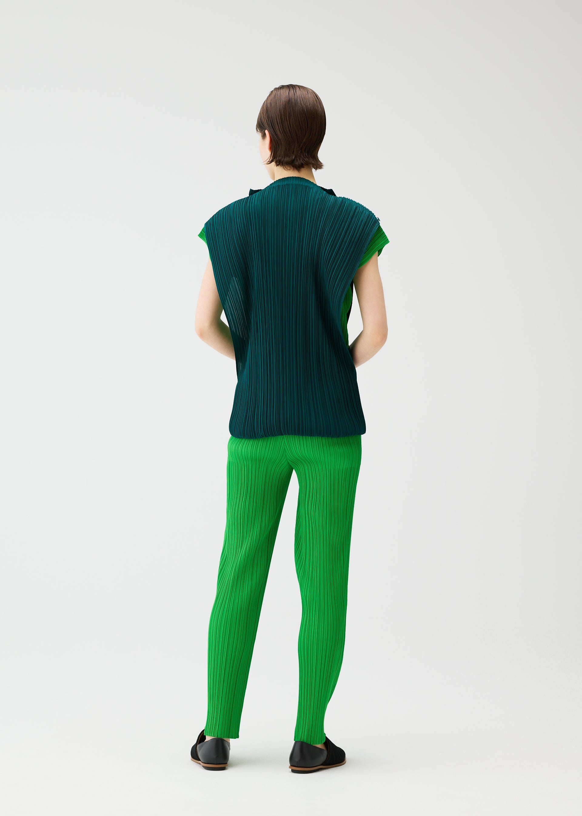 Zoomed view A person with short brown hair, facing away, wears PLEATS PLEASE ISSEY MIYAKE's SWING set: a dark green French-sleeve pleated top and bright green pleated pants with black shoes, set against a plain light background.
