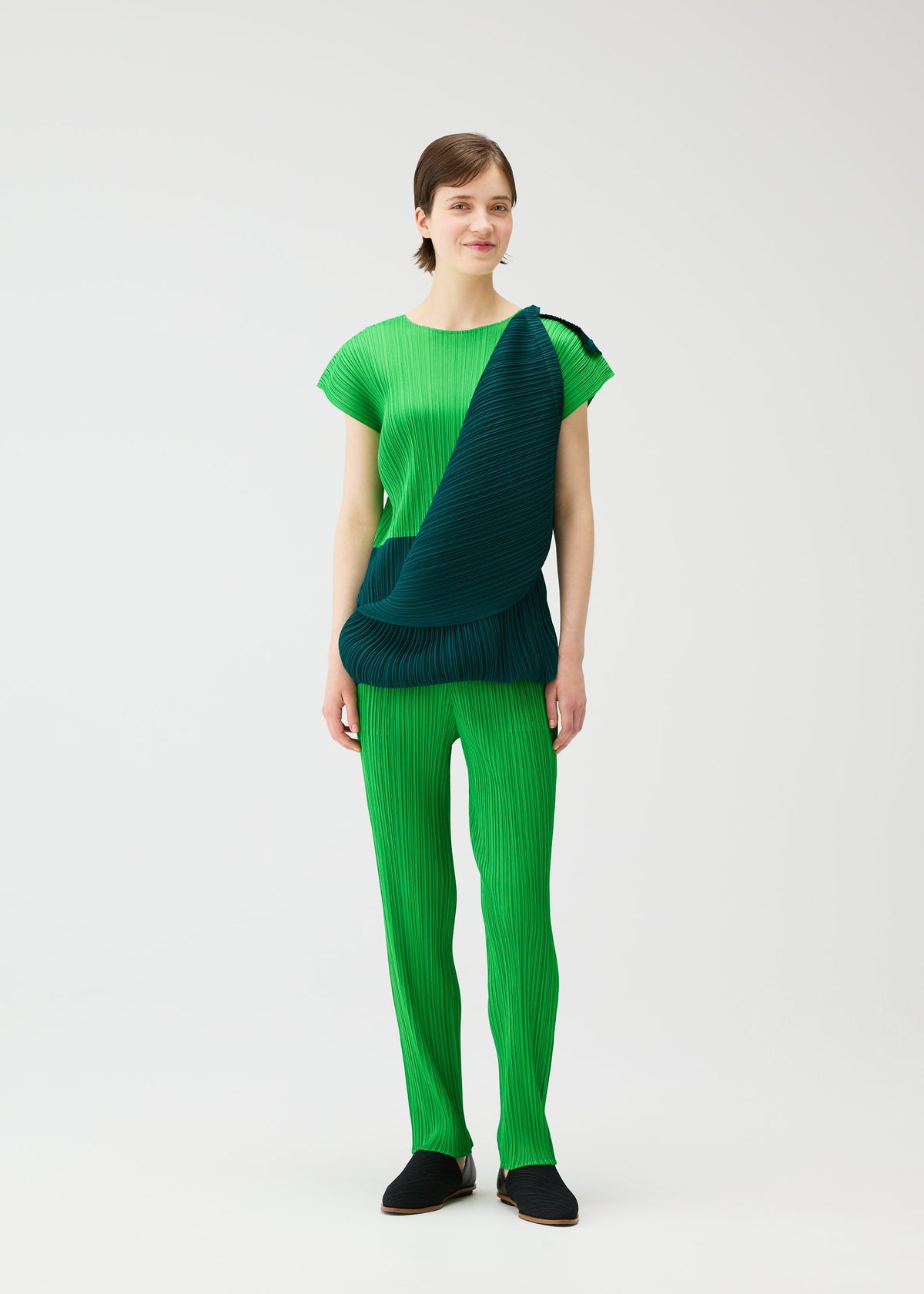 A woman with short brown hair smiles slightly as she models the SWING set by PLEATS PLEASE ISSEY MIYAKE: a bright green pleated French-sleeve top and pants, paired with a dark green pleated scarf draped diagonally, against a plain white background. view 13 of 14