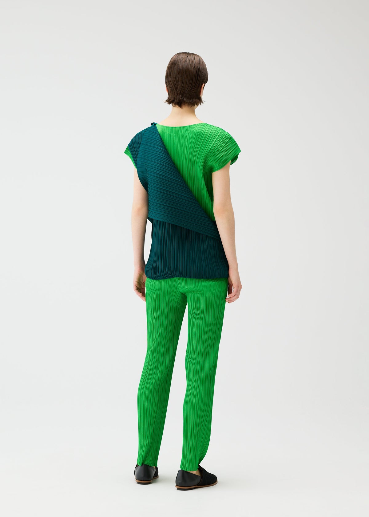 A person stands facing away, wearing the SWING dress by PLEATS PLEASE ISSEY MIYAKE—a bright green pleated, round-neck dress with French sleeves and a dark teal draped panel. A plain light background highlights its elegant two-tone design. view 14 of 14