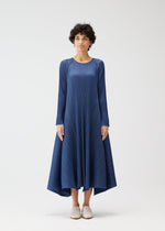 A person with short curly hair wears the CITY FRAME mid-calf blue pleated dress by PLEATS PLEASE ISSEY MIYAKE, featuring long sleeves and an asymmetrical hem, paired with light slip-on shoes against a plain white background.