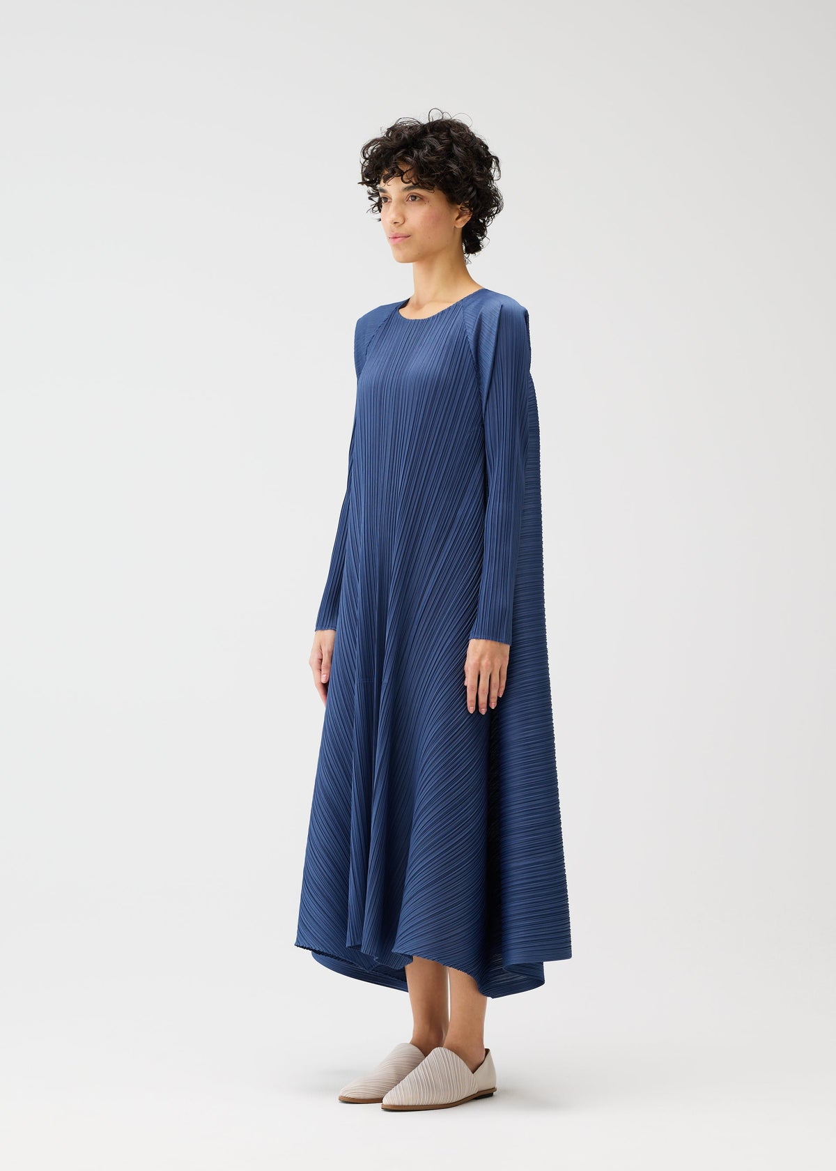 A woman with short curly hair wears the CITY FRAME blue long-sleeved pleated dress by PLEATS PLEASE ISSEY MIYAKE, featuring an asymmetrical hem and voluminous silhouette, paired with beige slip-on shoes against a plain white background. view 6 of 8