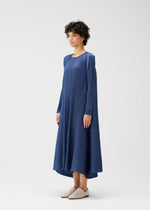 A woman with short curly hair wears the CITY FRAME blue long-sleeved pleated dress by PLEATS PLEASE ISSEY MIYAKE, featuring an asymmetrical hem and voluminous silhouette, paired with beige slip-on shoes against a plain white background.