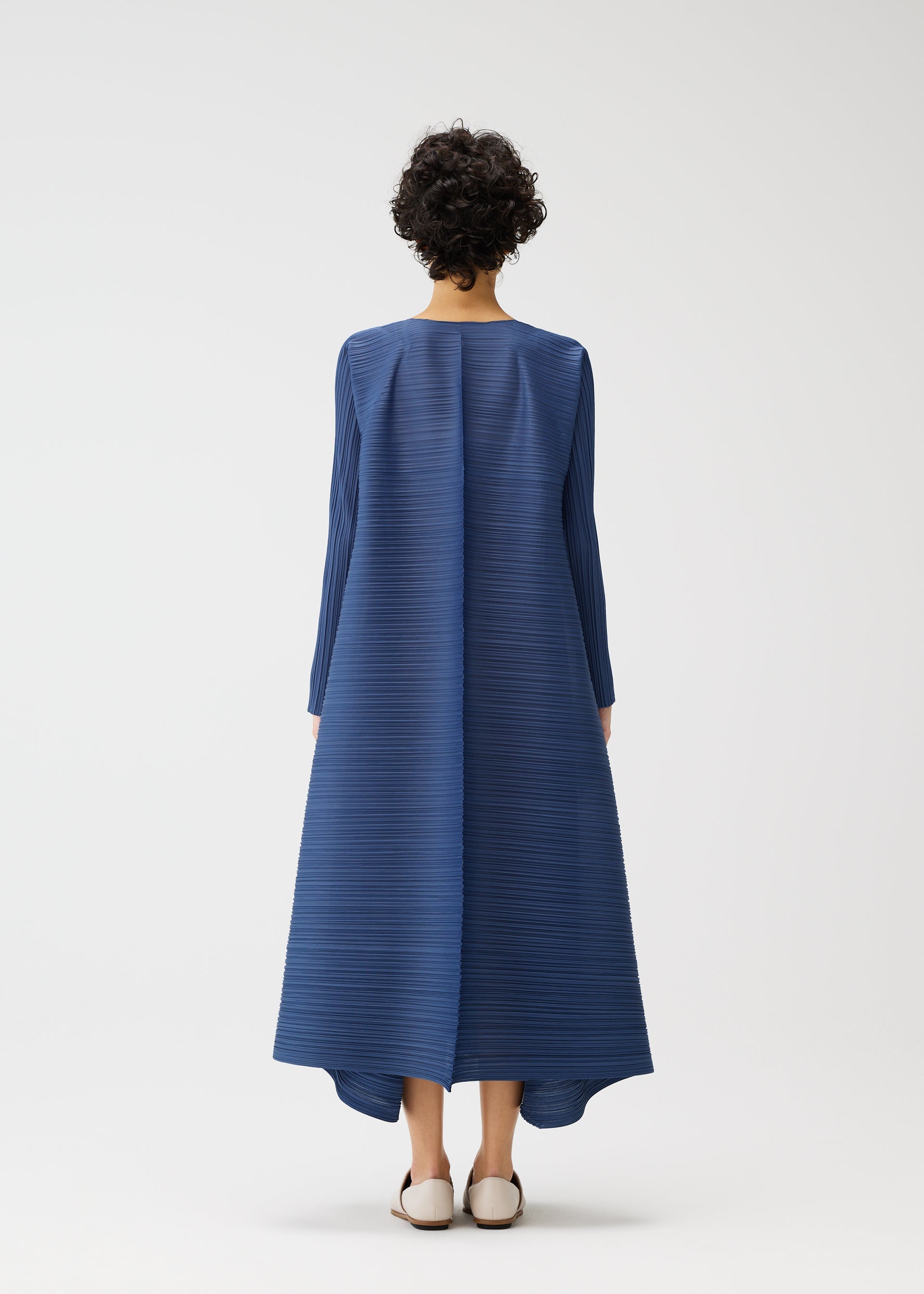 Zoomed view A person with short curly hair faces away, wearing the voluminous CITY FRAME blue dress with long sleeves by PLEATS PLEASE ISSEY MIYAKE. Light flat shoes complete the look against a plain white background.
