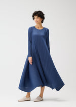 A person wears the CITY FRAME long-sleeved, round-neck pleated navy blue dress by PLEATS PLEASE ISSEY MIYAKE and light slip-on shoes, standing against a plain white background in a relaxed pose, looking off to the side.