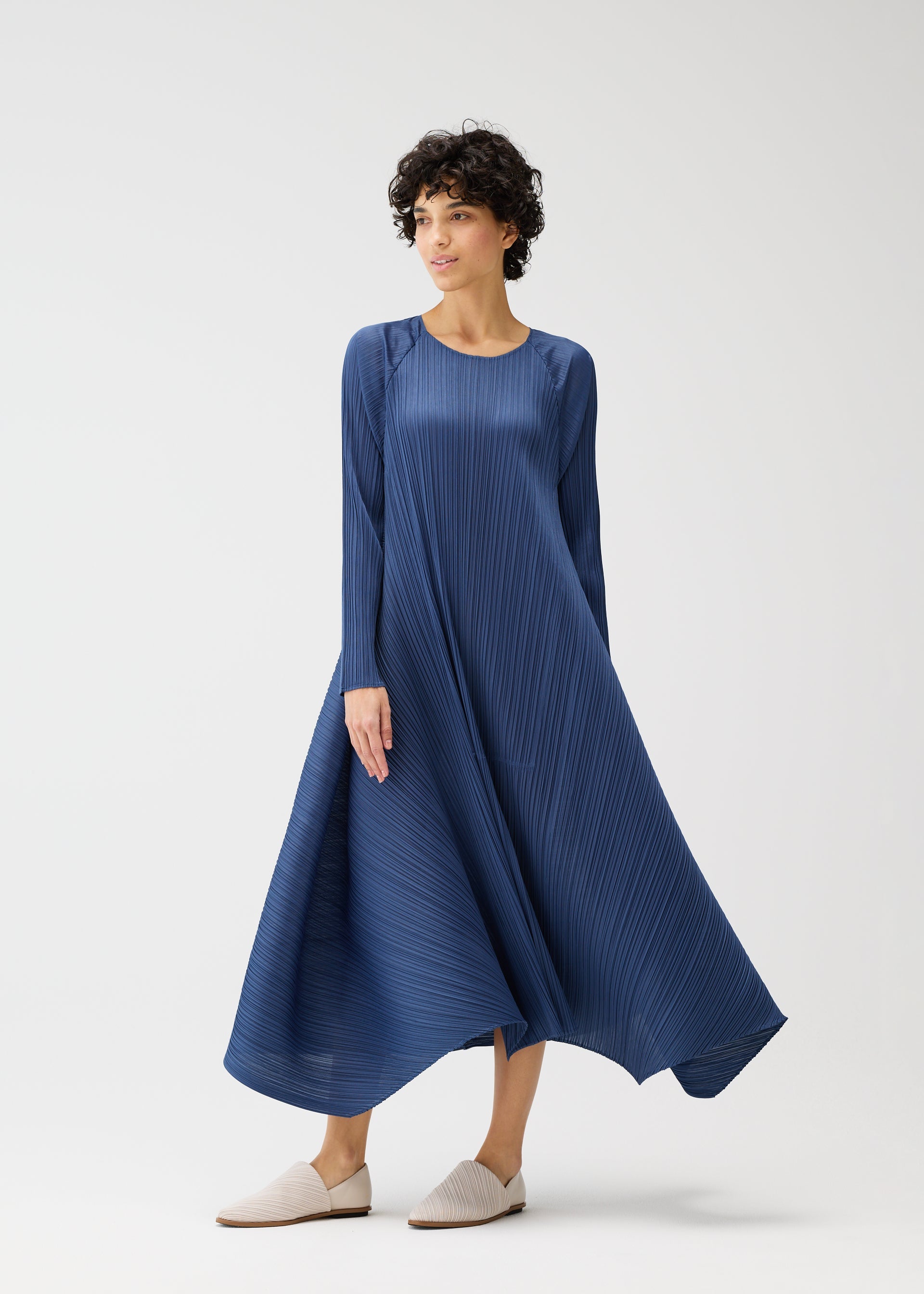 Zoomed view A person wears the CITY FRAME long-sleeved, round-neck pleated navy blue dress by PLEATS PLEASE ISSEY MIYAKE and light slip-on shoes, standing against a plain white background in a relaxed pose, looking off to the side.