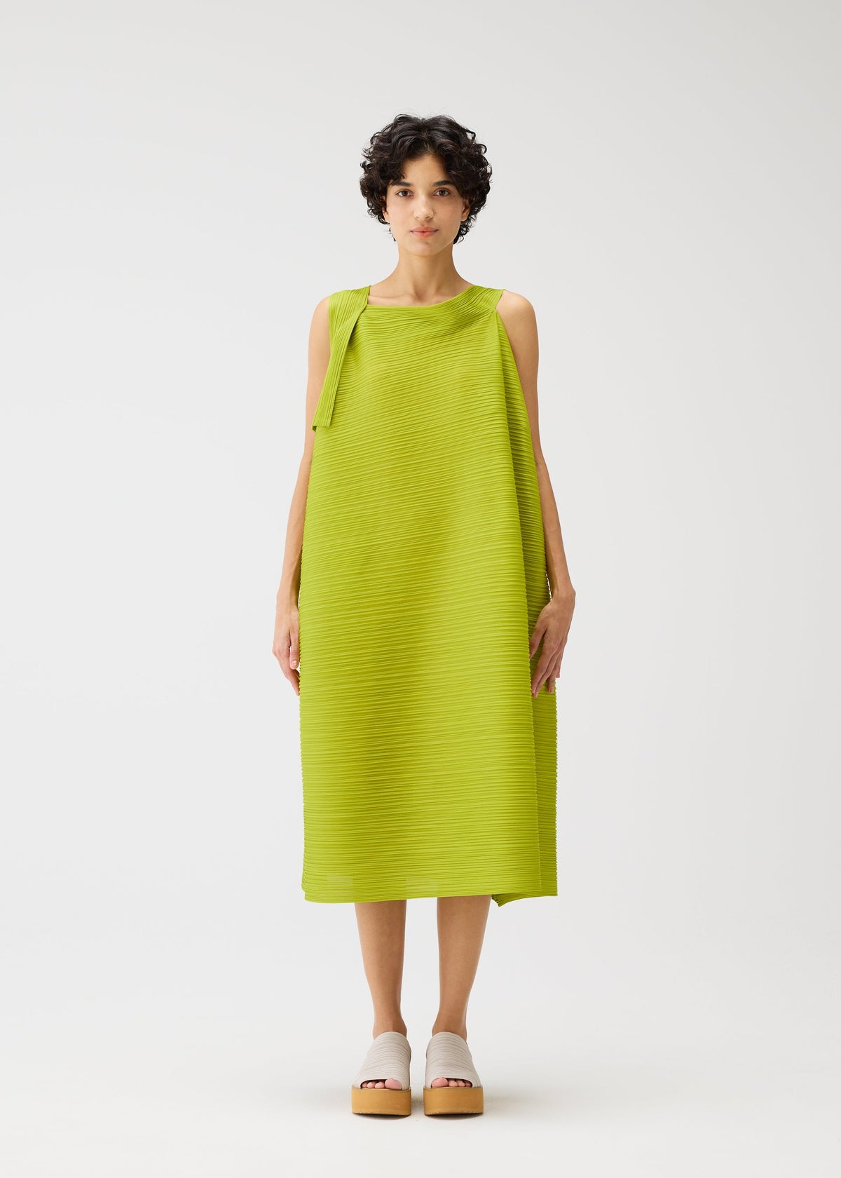 Against a plain white background, a person with short curly hair wears the BOUNCY BASKET dress by PLEATS PLEASE ISSEY MIYAKE—a sleeveless, bright green, textured midi dress—paired with beige platform sandals. view 5 of 8