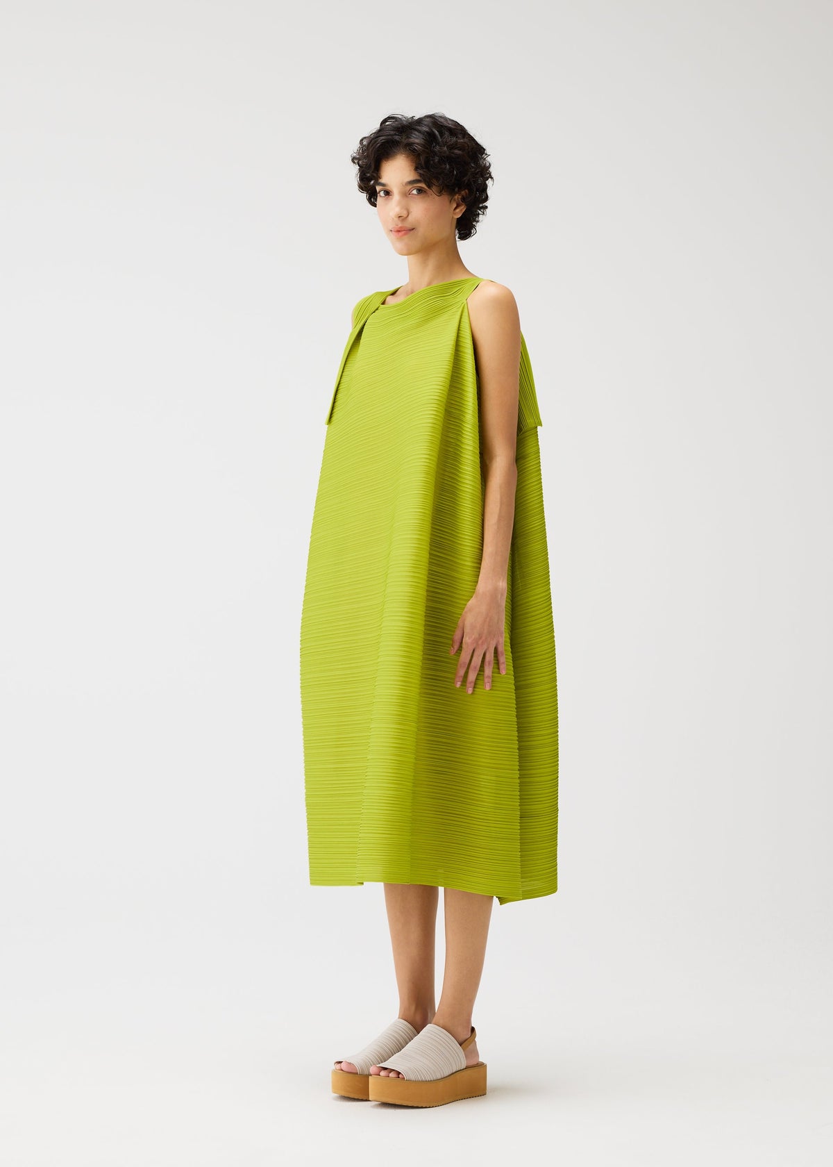 A person with short curly hair stands against a white background, wearing the BOUNCY BASKET sleeveless dress by PLEATS PLEASE ISSEY MIYAKE in bright green, paired with light-colored platform sandals. view 6 of 8