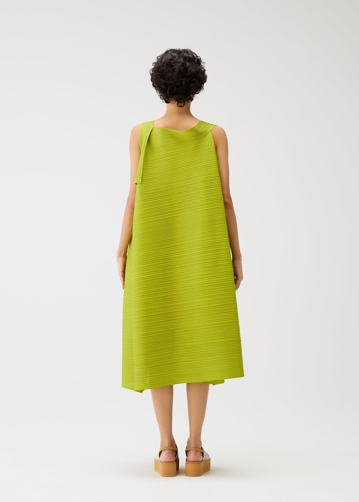 A person with short curly hair faces away, wearing the BOUNCY BASKET dress by PLEATS PLEASE ISSEY MIYAKE—lime green, sleeveless, basket-inspired with an asymmetrical drape—and tan platform sandals, against a plain white background. view 7 of 8