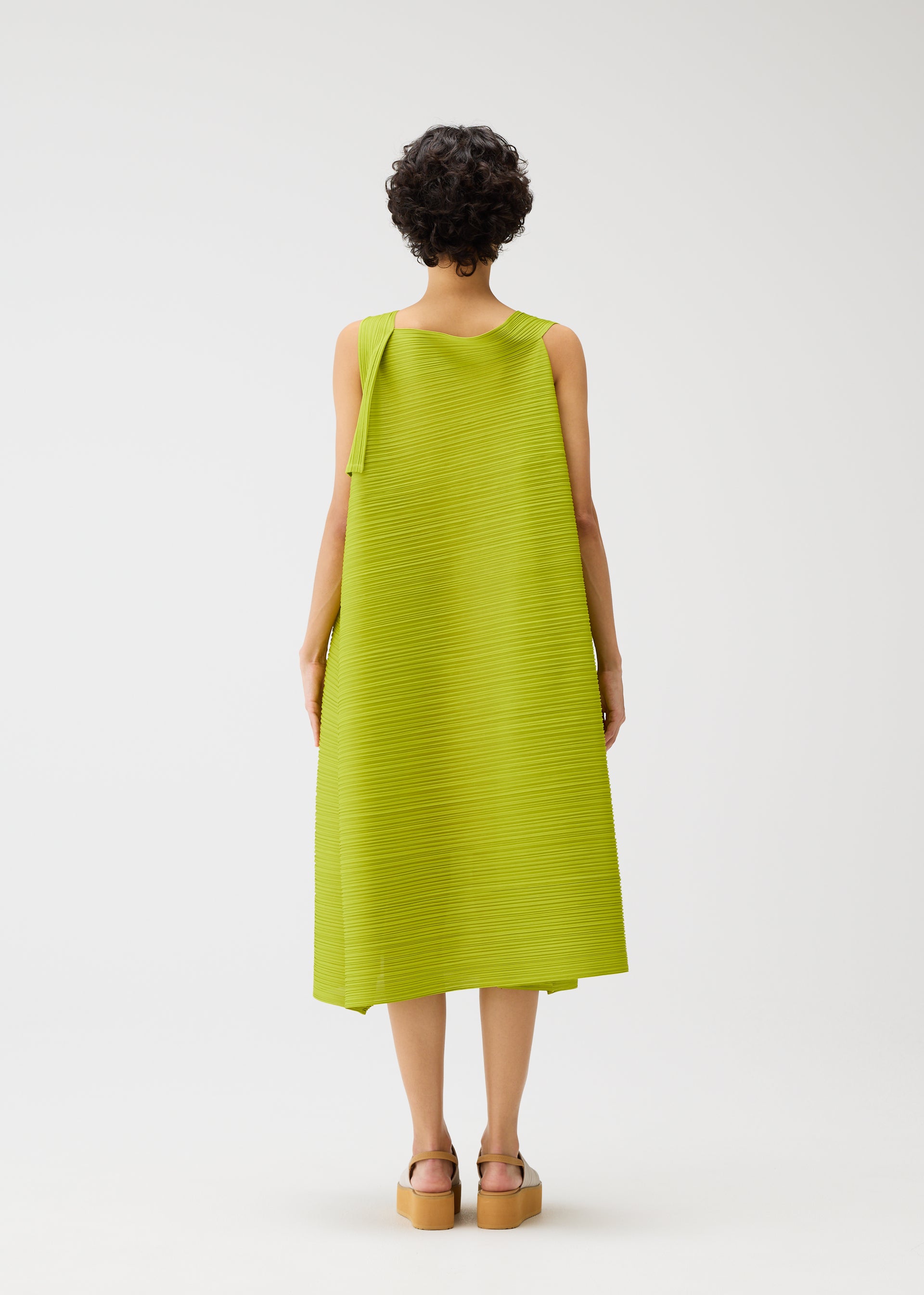 Zoomed view A person with short curly hair faces away, wearing the BOUNCY BASKET dress by PLEATS PLEASE ISSEY MIYAKE—lime green, sleeveless, basket-inspired with an asymmetrical drape—and tan platform sandals, against a plain white background.
