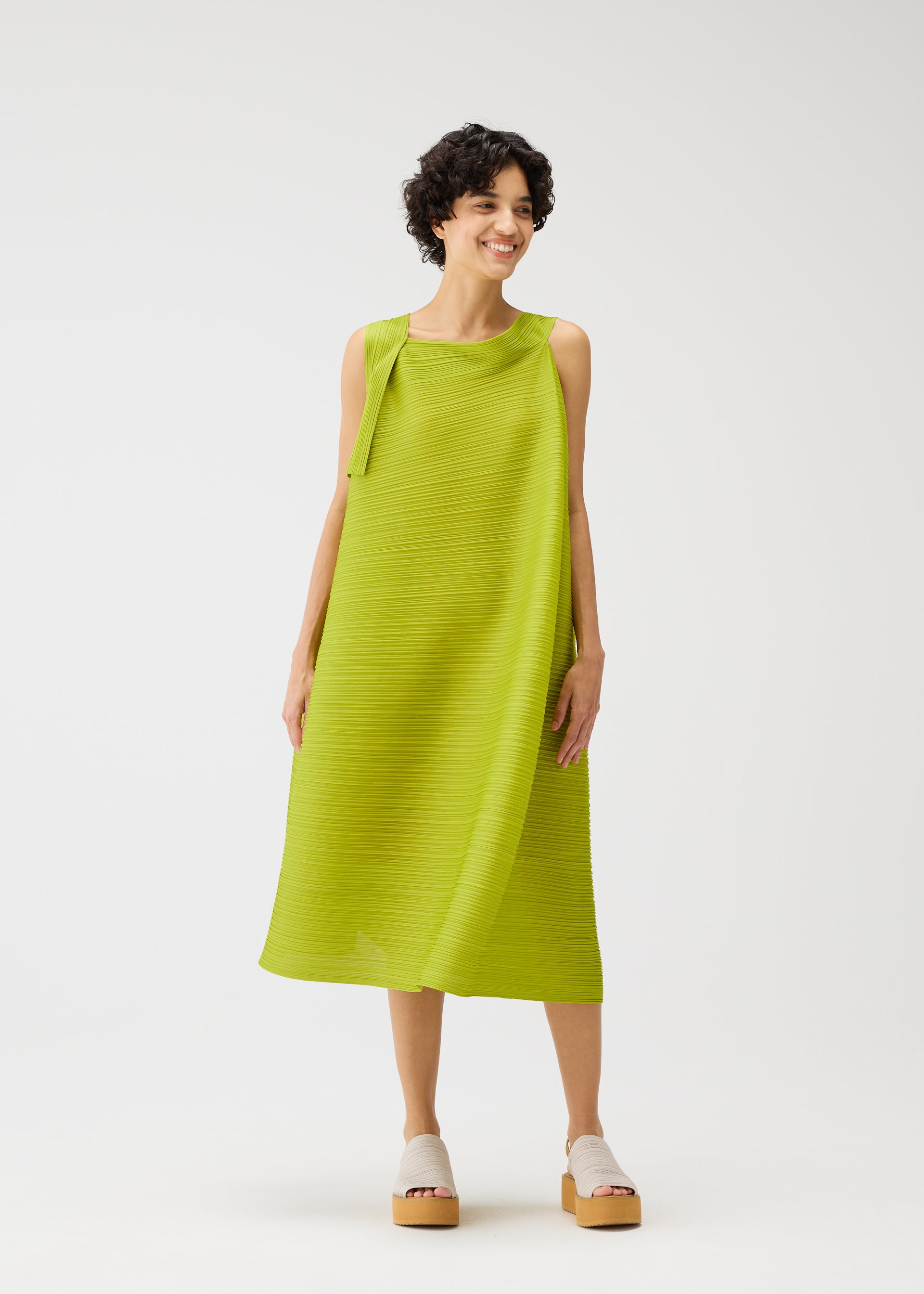 Zoomed view A person with short dark hair smiles while wearing the lime green BOUNCY BASKET pleated midi dress by PLEATS PLEASE ISSEY MIYAKE and light platform sandals, standing against a plain white background.