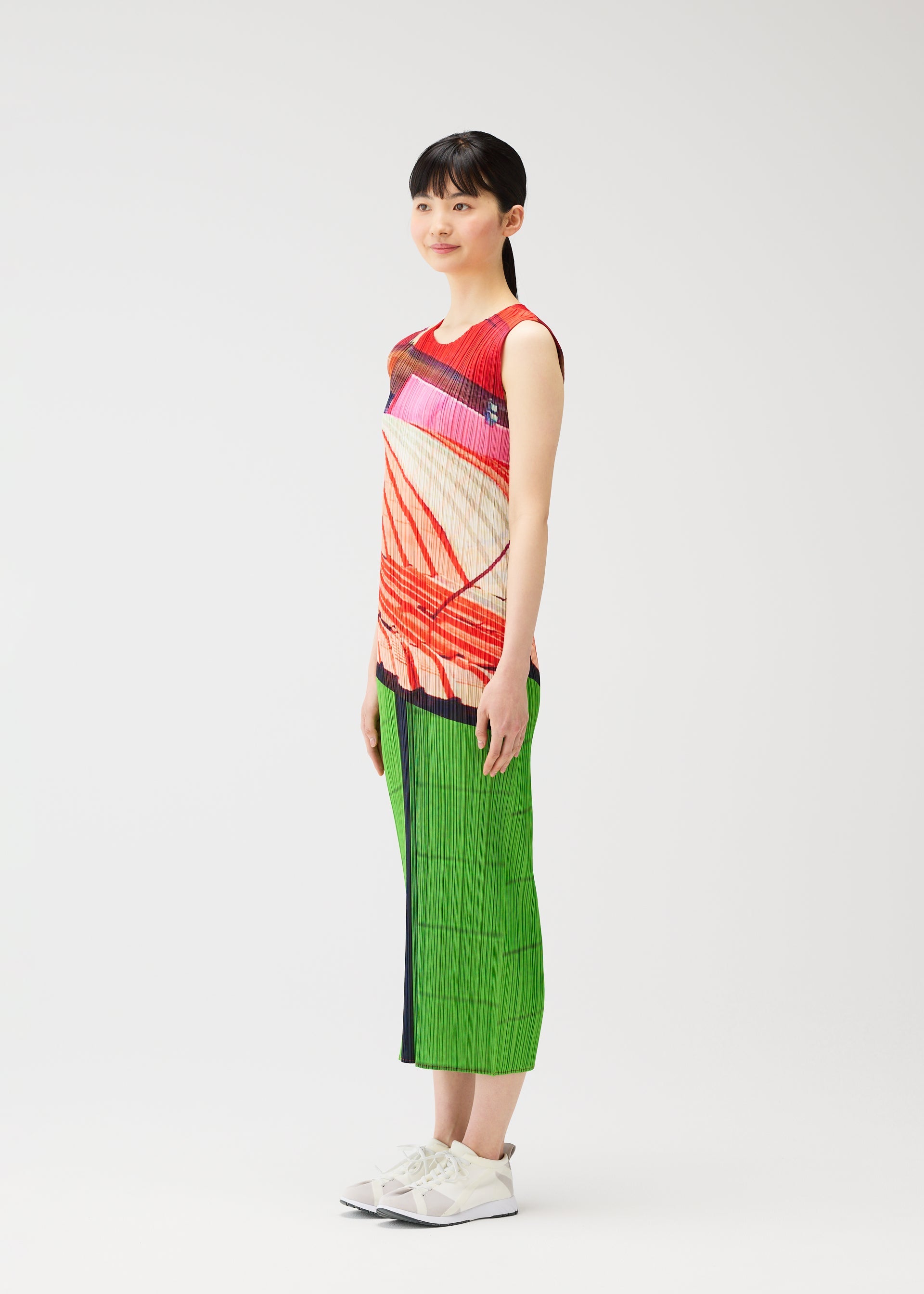 Zoomed view A woman with straight dark hair wears the CITY COLLAGE sleeveless top by PLEATS PLEASE ISSEY MIYAKE, featuring red and white collage patterns, paired with green pleated pants and white sneakers in a studio with a plain white background.