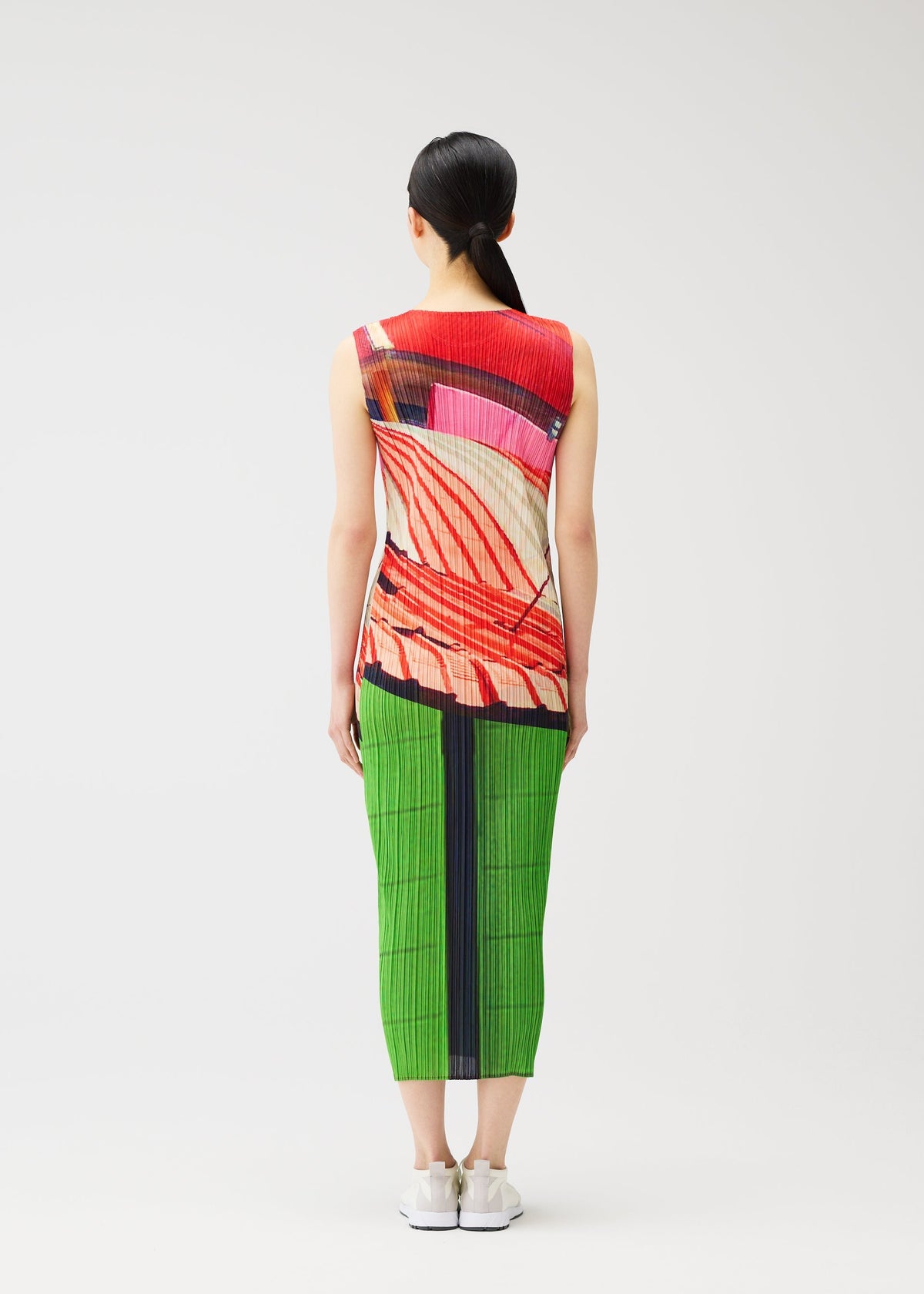 A person with long dark hair in a low ponytail stands facing away, wearing the CITY COLLAGE sleeveless dress by PLEATS PLEASE ISSEY MIYAKE, featuring red, pink, and beige collage patterns and a bright green skirt. The background is plain white. view 7 of 7