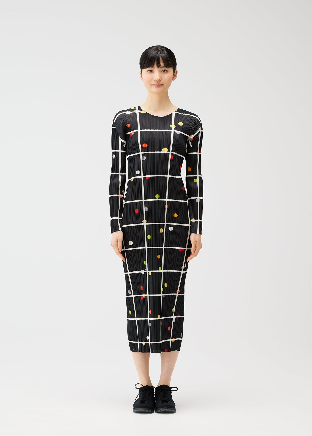 A woman faces forward wearing the PLEATS PLEASE ISSEY MIYAKE CROSSING DOTS dress, a long-sleeved, form-fitting round-neck style with a black base, white grid pattern, and colorful dots. She has straight black hair and wears black shoes against a white background. view 4 of 7