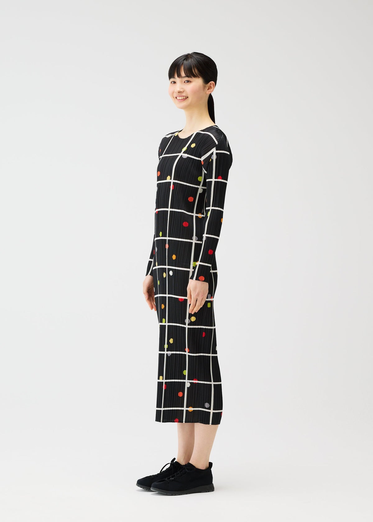 A woman with straight dark hair and bangs smiles sideways, modeling the ankle-length CROSSING DOTS dress by PLEATS PLEASE ISSEY MIYAKE—black with a white grid and colorful dots. She pairs it with black sneakers against a plain white background. view 5 of 7