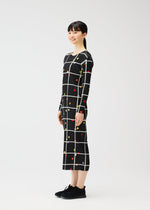 A woman with straight dark hair and bangs smiles sideways, modeling the ankle-length CROSSING DOTS dress by PLEATS PLEASE ISSEY MIYAKE—black with a white grid and colorful dots. She pairs it with black sneakers against a plain white background.