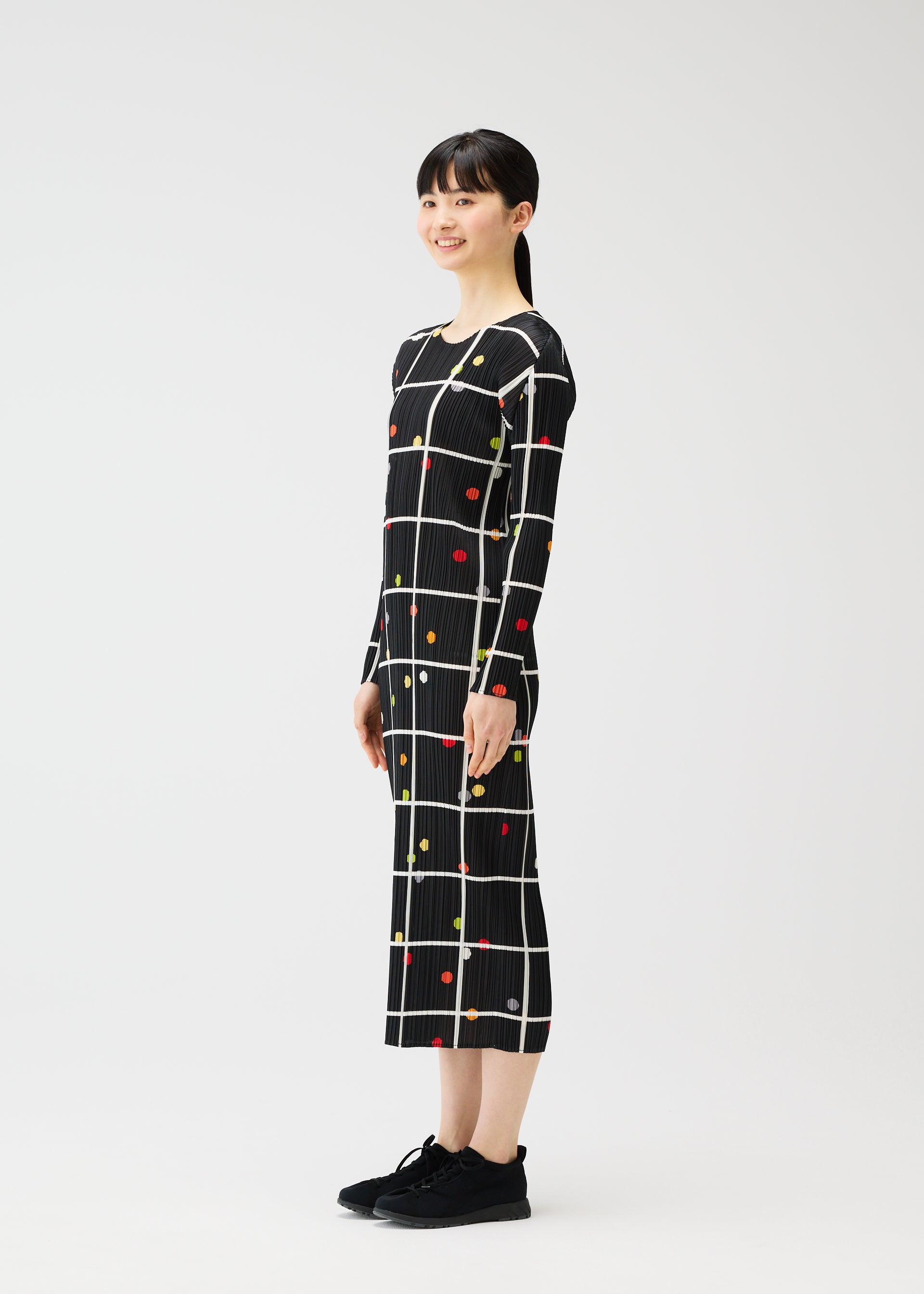Zoomed view A woman with straight dark hair and bangs smiles sideways, modeling the ankle-length CROSSING DOTS dress by PLEATS PLEASE ISSEY MIYAKE—black with a white grid and colorful dots. She pairs it with black sneakers against a plain white background.