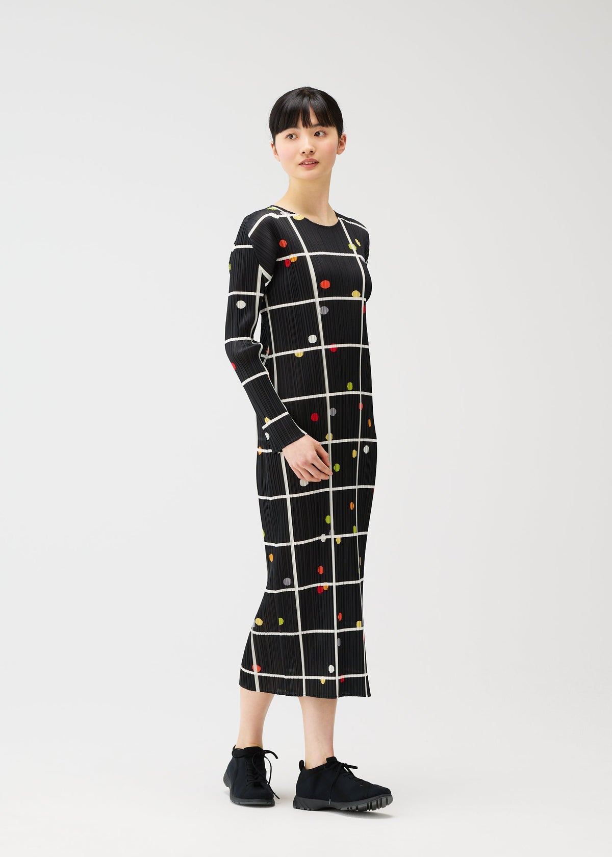A woman stands against a white background wearing the CROSSING DOTS dress by PLEATS PLEASE ISSEY MIYAKE, a round-neck, long-sleeved piece with a black grid and colorful dots pattern, paired with black sneakers. view 7 of 7