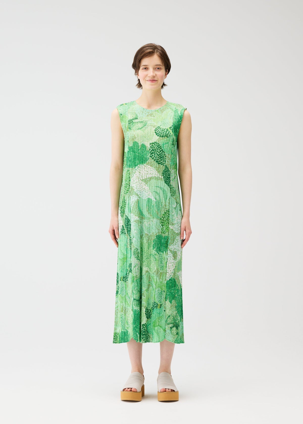 A woman with short brown hair faces forward in PLEATS PLEASE ISSEY MIYAKE's FOGGY FOREST, a sleeveless green and white flared dress with pleated texture and pointillism gradation. She wears light sandals against a plain white background. view 4 of 7